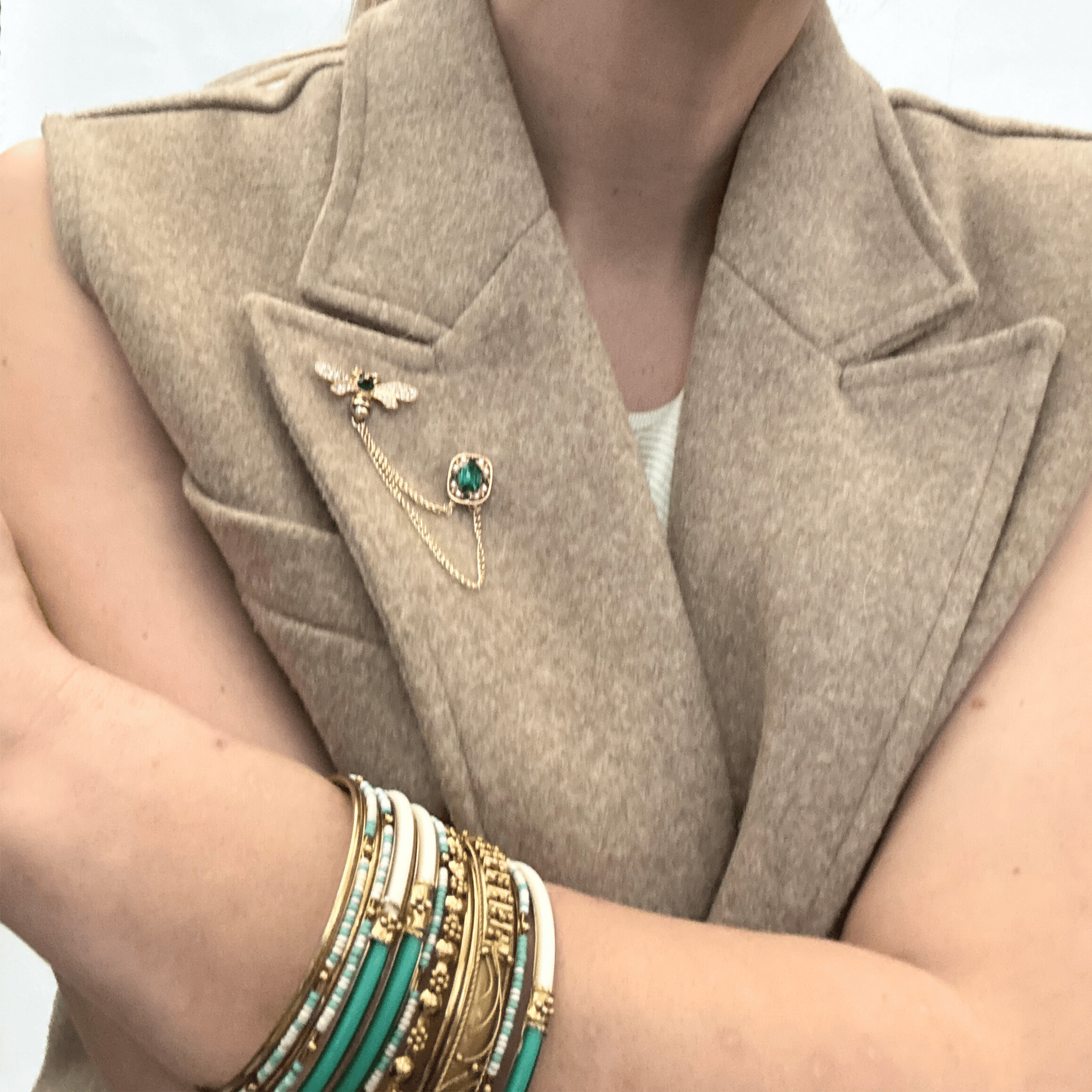 A brilliant, emerald-toned teardrop stone set in a textured gold-tone frame, flanked by delicate crystal-encrusted wings, styled on the lapel of a sleeves wool vest.
