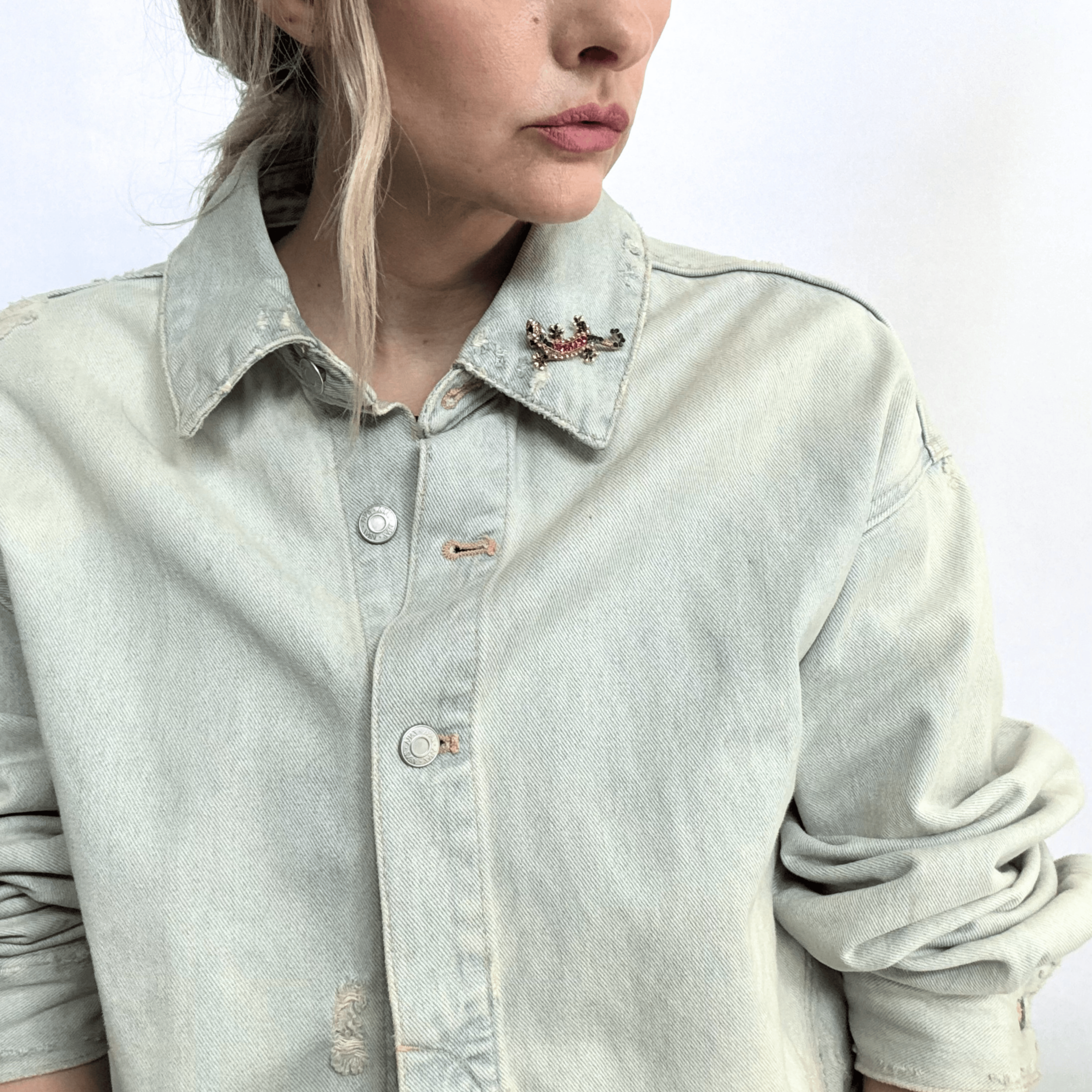 The Kaleidoscope Climber Brooch is a jewel toned gecko shaped brooch in a gold toned setting, styled on the tip of the collar of a denim jacket.