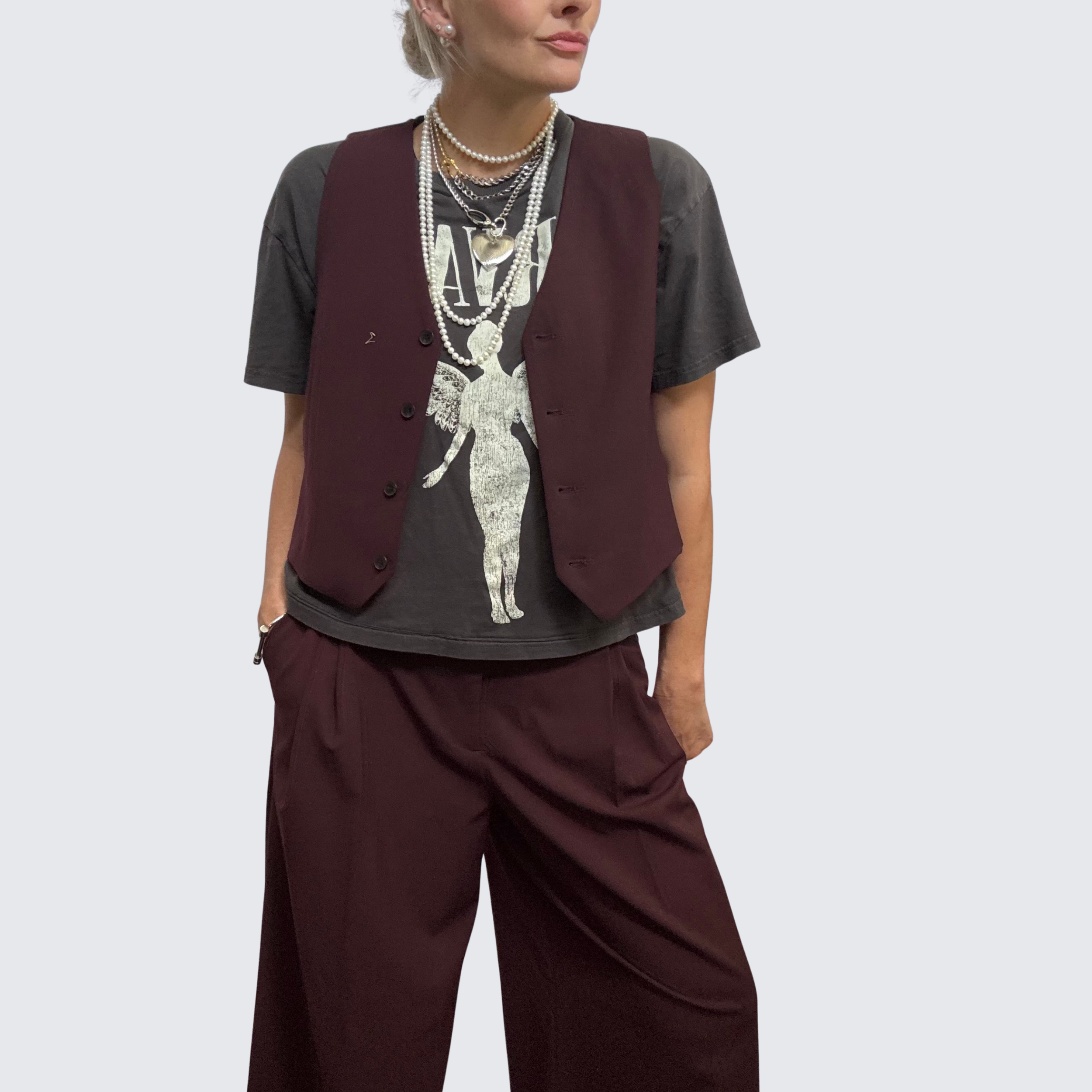 Woman wearing plum vest and trousers with a graphic t-shirt. She wears a bold necklace stack featuring multiple pearl strands and chunky silver chains from The Brooch House.