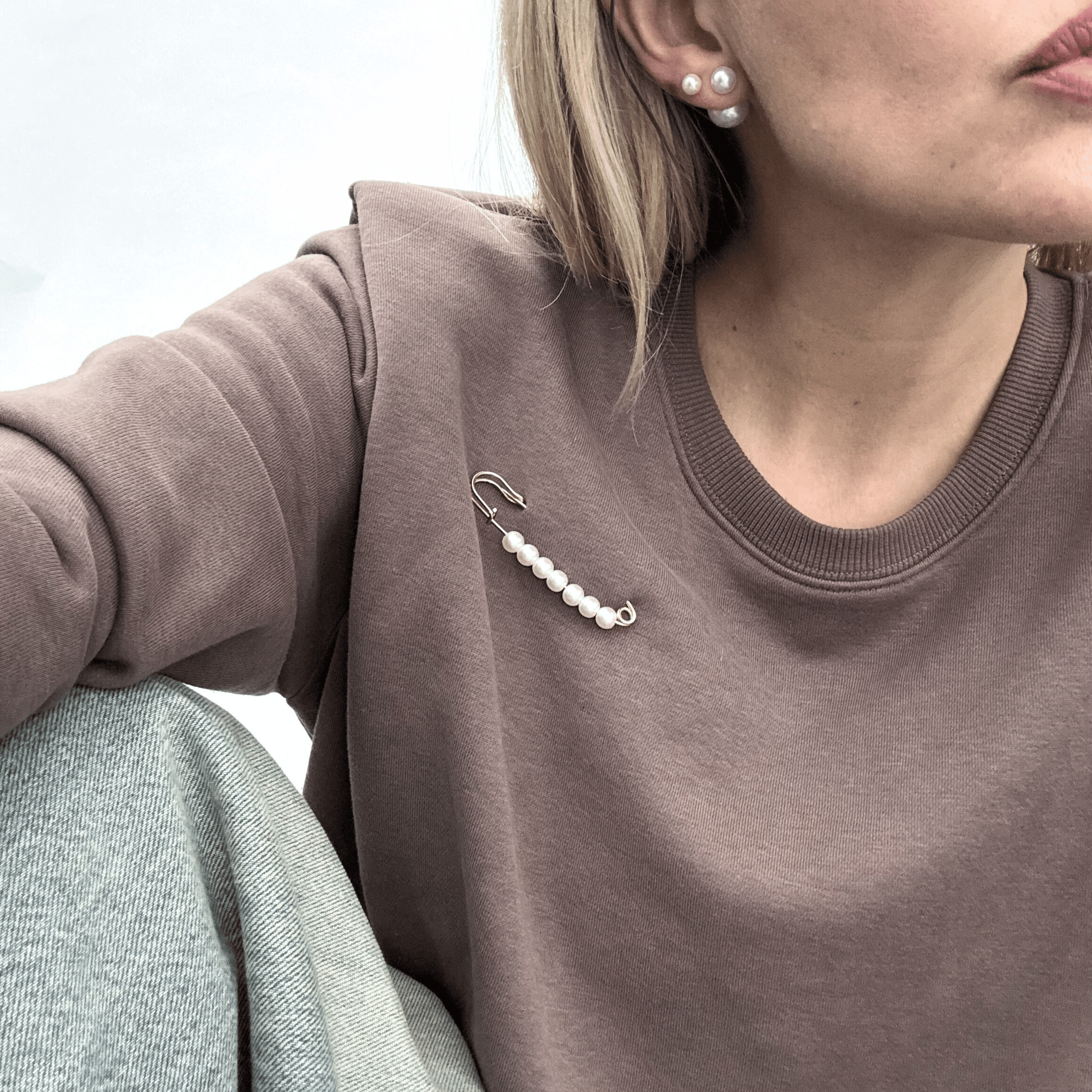 Gold toned linear safety pin, featuring a row of five graduated, high-lustre faux pearls, pinned on a brown sweatshirt.