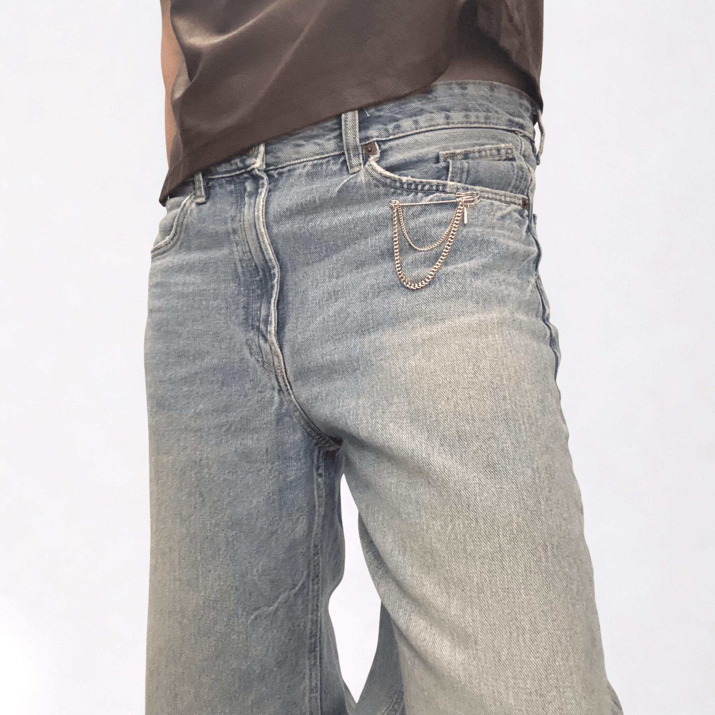 Golden oversized safety pin brooch with a double-tier of delicate curb chains, styled on the pocket of a pair of oversized jeans.