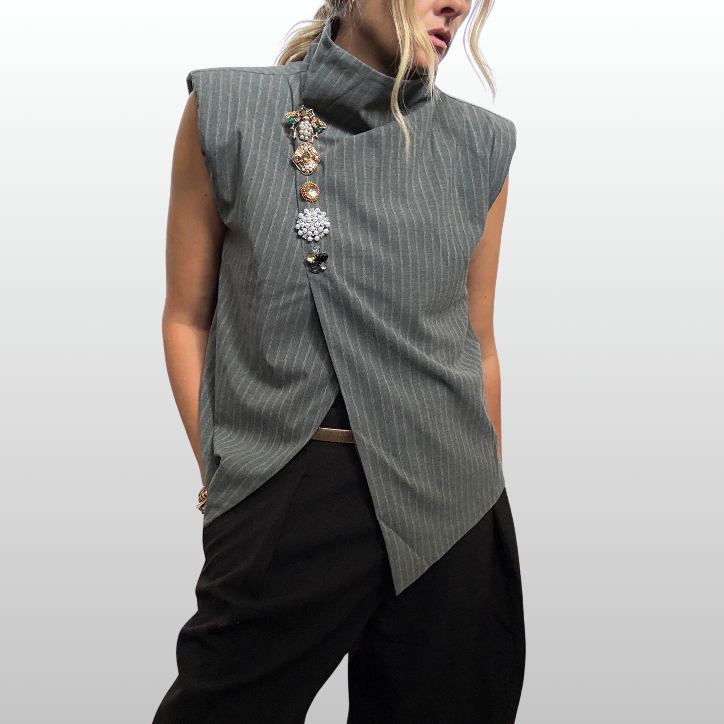 Close up of woman wearing an asymmetric sleeves grey pin striped vest. Ar the closure of the vest is a vertical stack of 5 brooches in various gold and amber tones.
