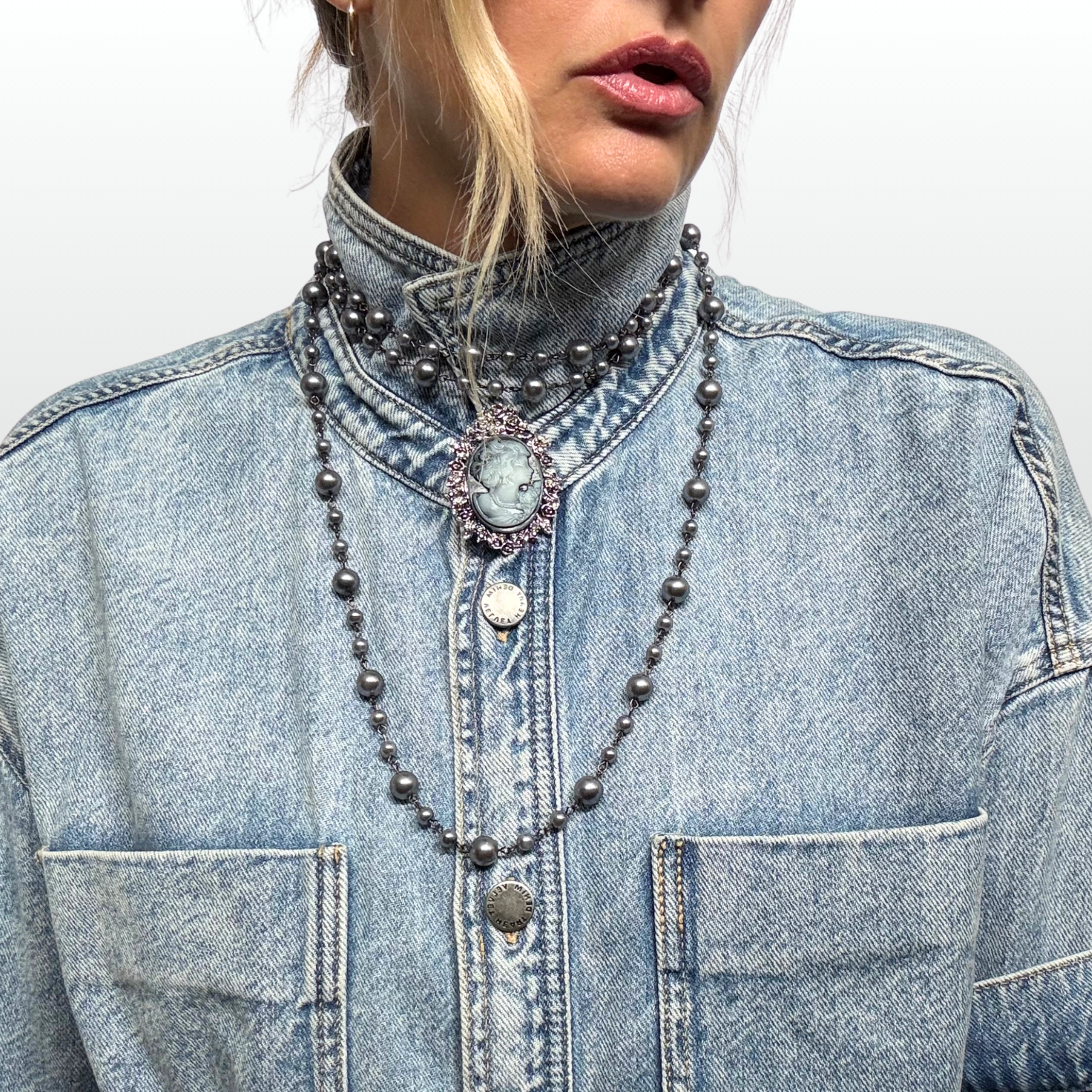 Woman wearing a denim short and multiple grey pearl strands. in the centre of her collar sits a grey blue cameo brooch.