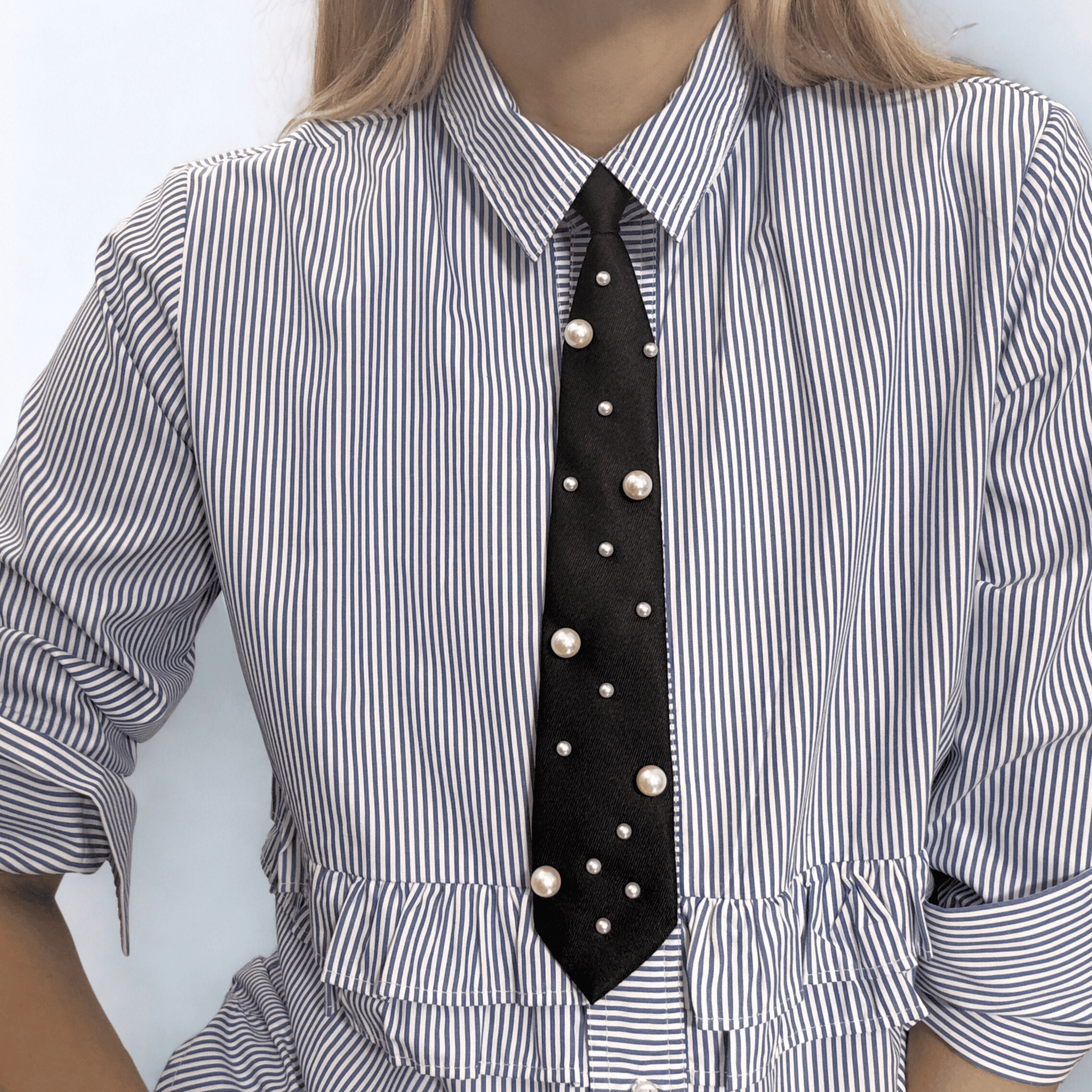 High-lustre faux pearls are scattered in an organic, constellation-like pattern across matte-black fabric neck tie.  Styled on a blue striped button up with ruffle detail.