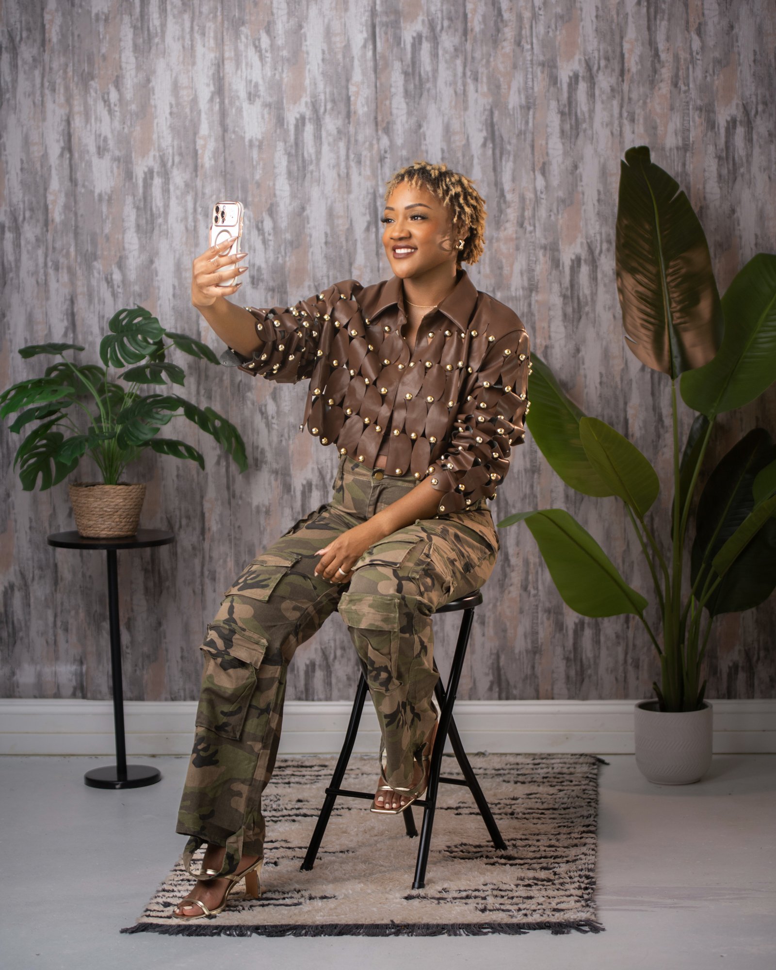 A woman with short curly hair, wearing a brown shirt with gold embellishments and camouflage cargo pants, sitting on a stool and taking a selfie with her phone. There are two potted plants and textured wallpaper in the background.