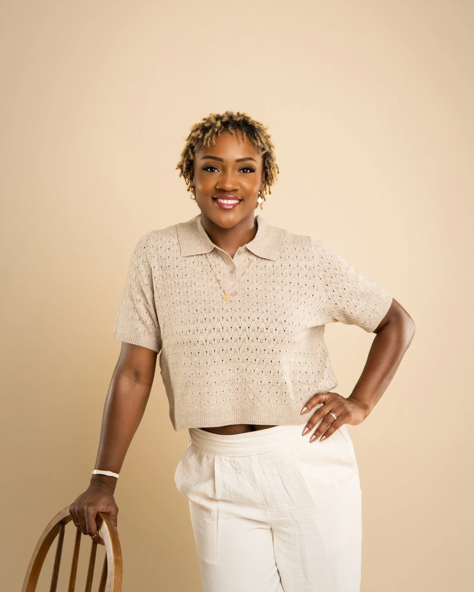 A smiling woman with short, curly blonde hair, wearing a beige knit polo shirt and cream-colored pants, standing against a neutral background with one hand on her hip and the other resting on a wooden chair.