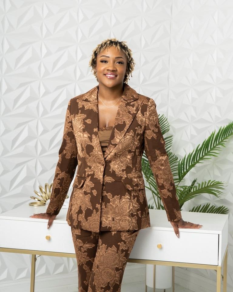 A woman in a brown floral blazer and matching pants standing behind a white console table with gold accents, smiling against a white textured wall and green plant.