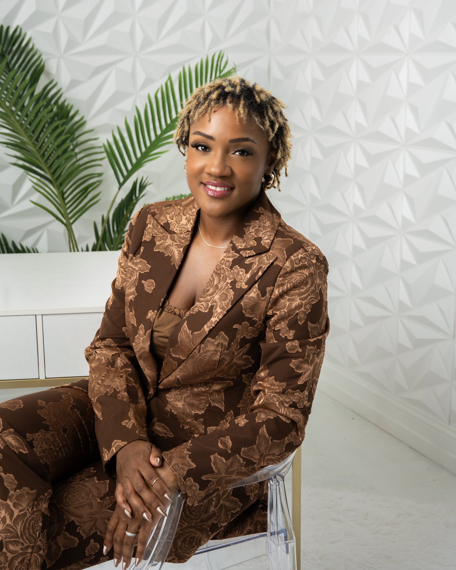 A woman with short blonde curly hair, wearing a brown floral patterned suit, sitting on a clear plastic chair in a modern room with white geometric wall and green plants behind her.