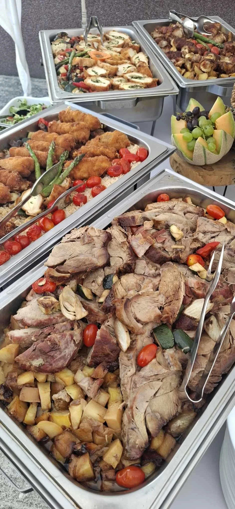 Buffet table with trays of roasted meats, vegetables, and stuffed chicken, along with a bowl of mixed fruit.