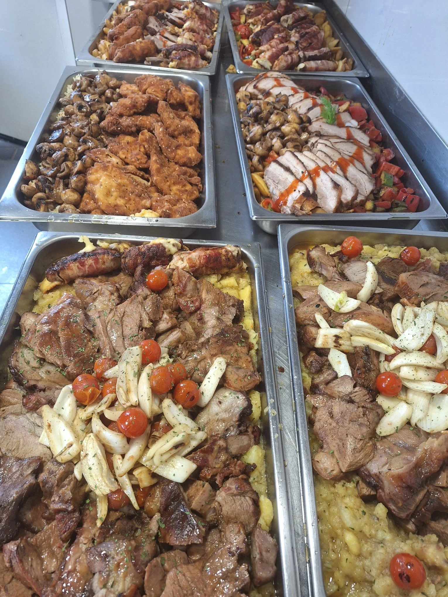 Several trays of cooked meats and vegetables, including roast beef, pork, fried chicken, chicken wings, mushrooms, roasted peppers, and garlic, on a stainless steel countertop.