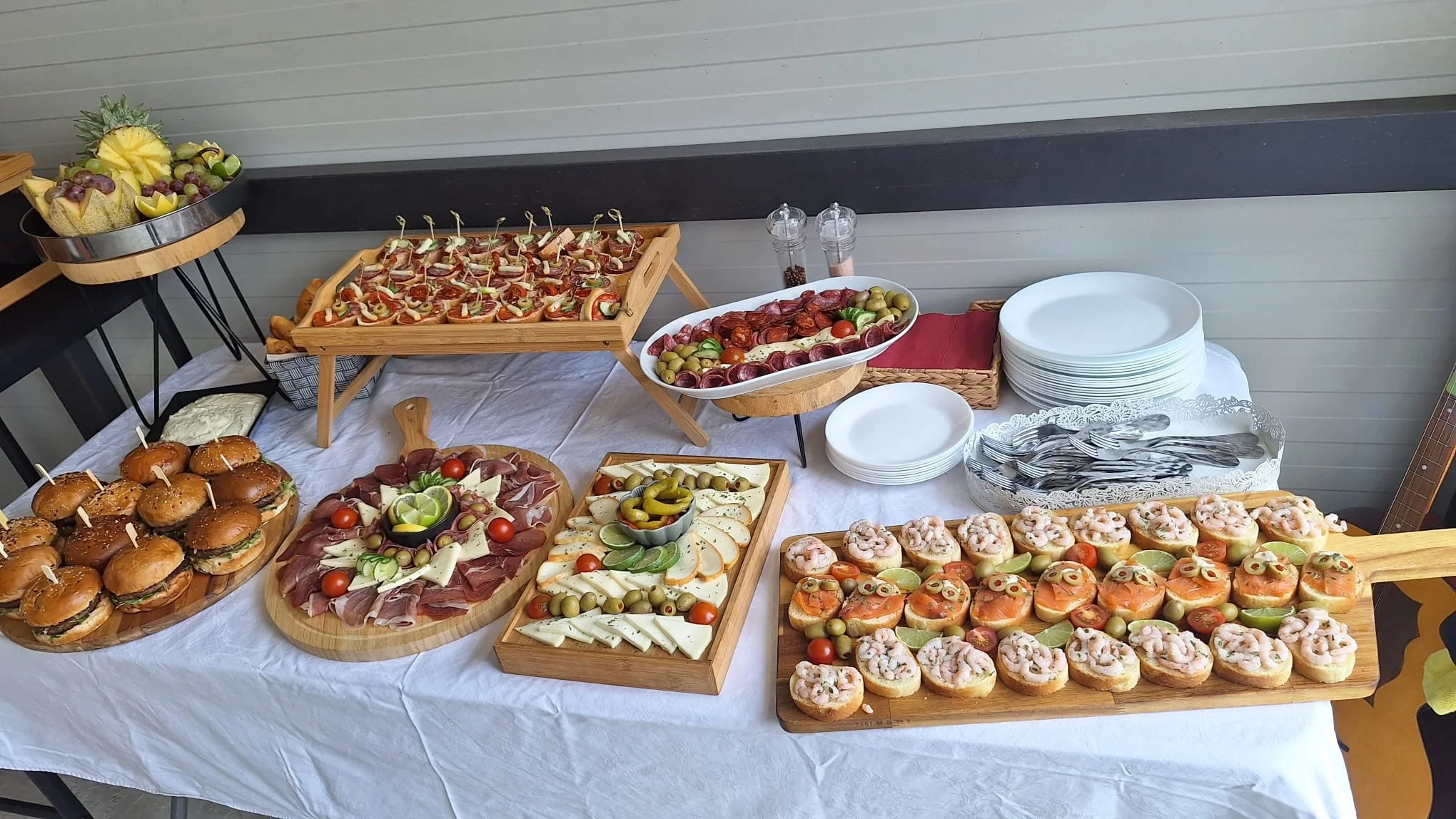 A table with a white tablecloth displays an assortment of finger foods including sliders, charcuterie, cheese, shrimp canapés, and various garnishes, along with stacked plates, forks, and condiments.