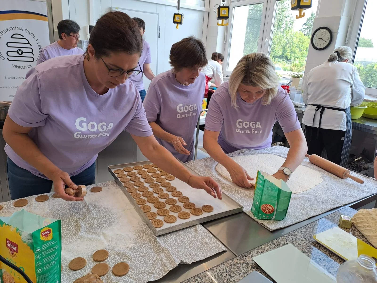 Three women in matching lavender shirts with 'GOGA Gluten Free' printed on them are baking cookies in a bright kitchen. They are rolling out dough and placing cookies on baking sheets, with ingredients and baking tools on the counter.