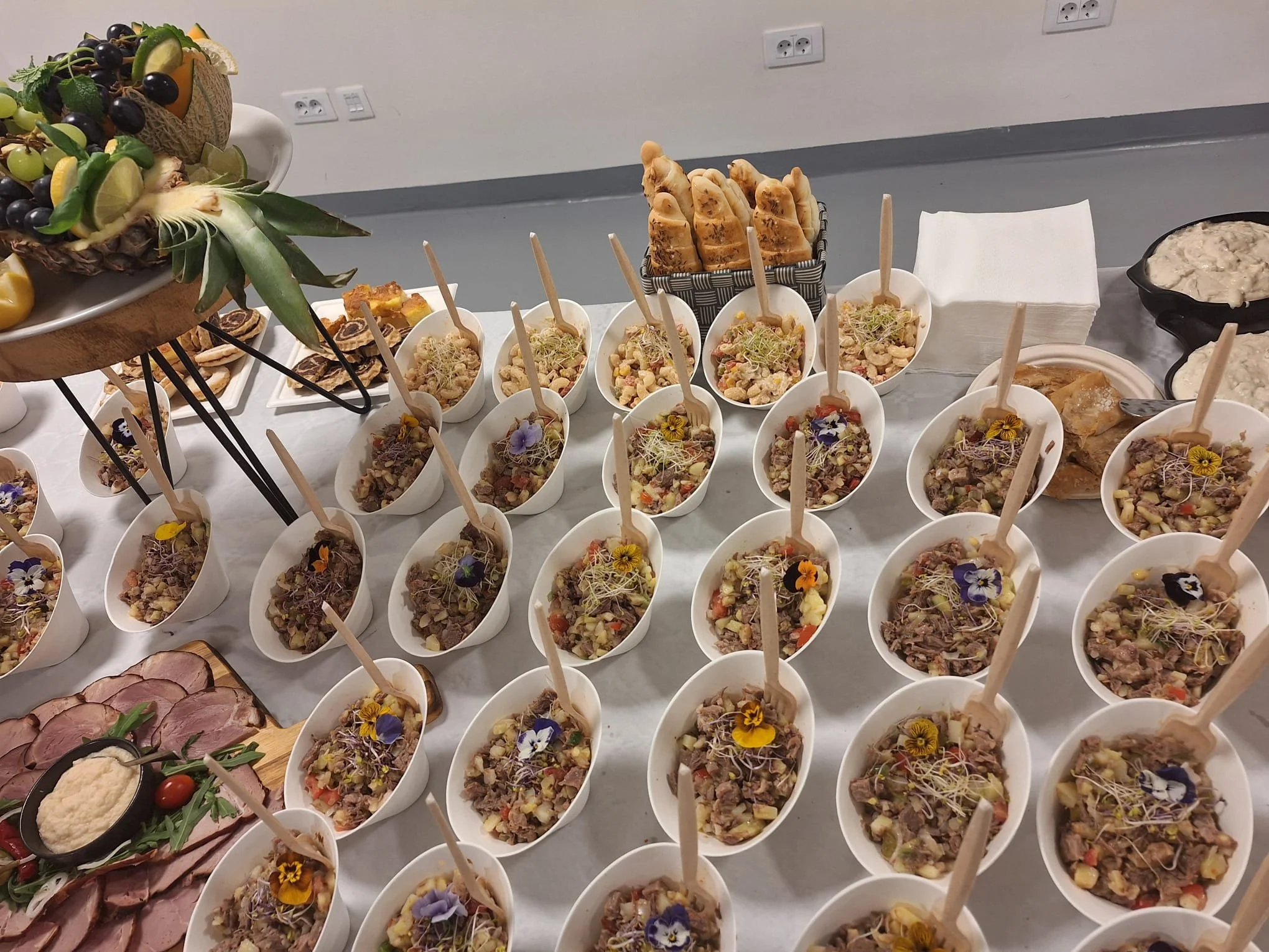 A table with multiple small bowls filled with a salad topped with edible flowers, with bread, sliced meats, and fruit arrangements in the background.