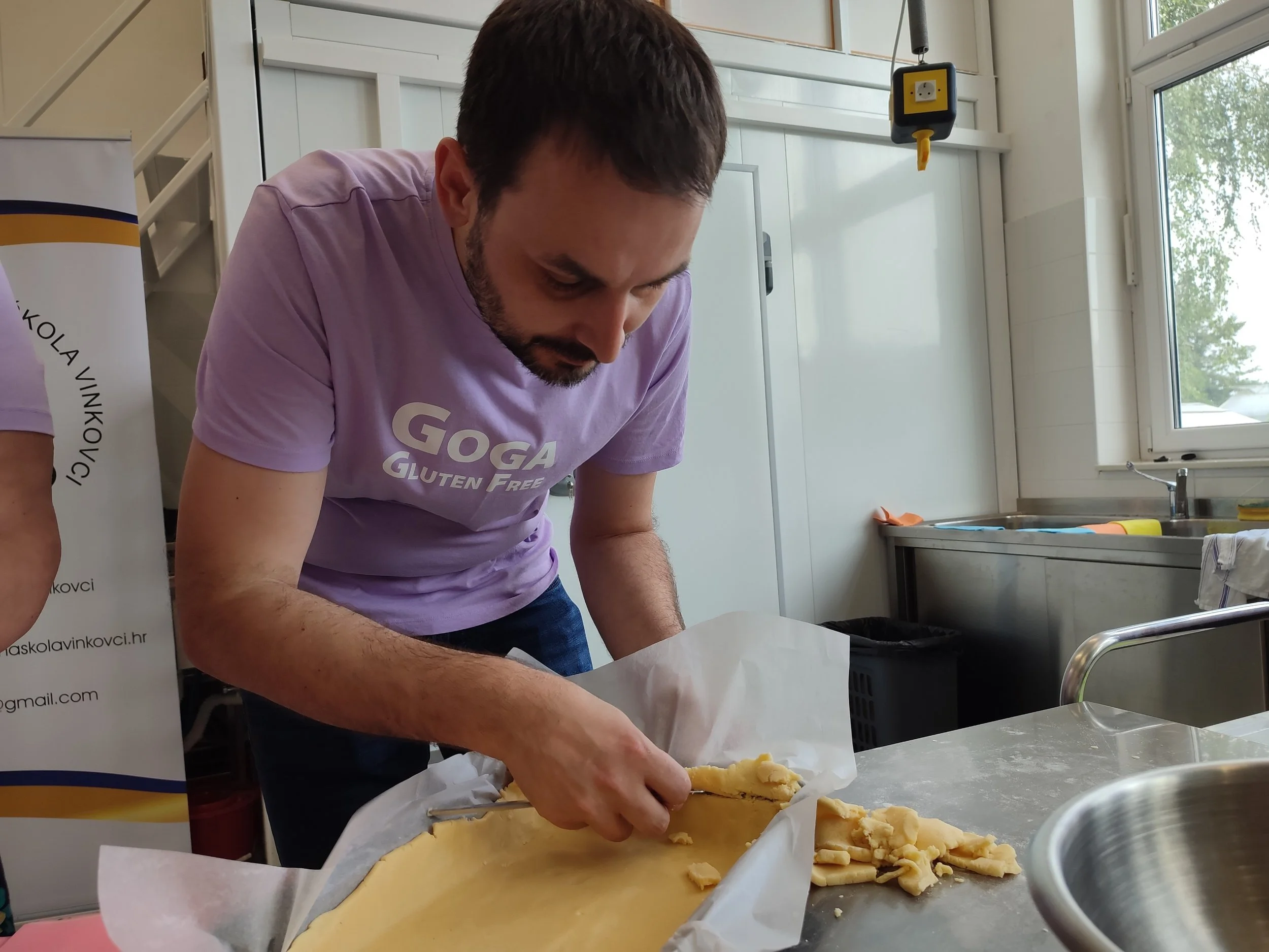 A man in a purple T-shirt with the text 'GOGA Gluten Free' is working with dough on a baking sheet in a kitchen.