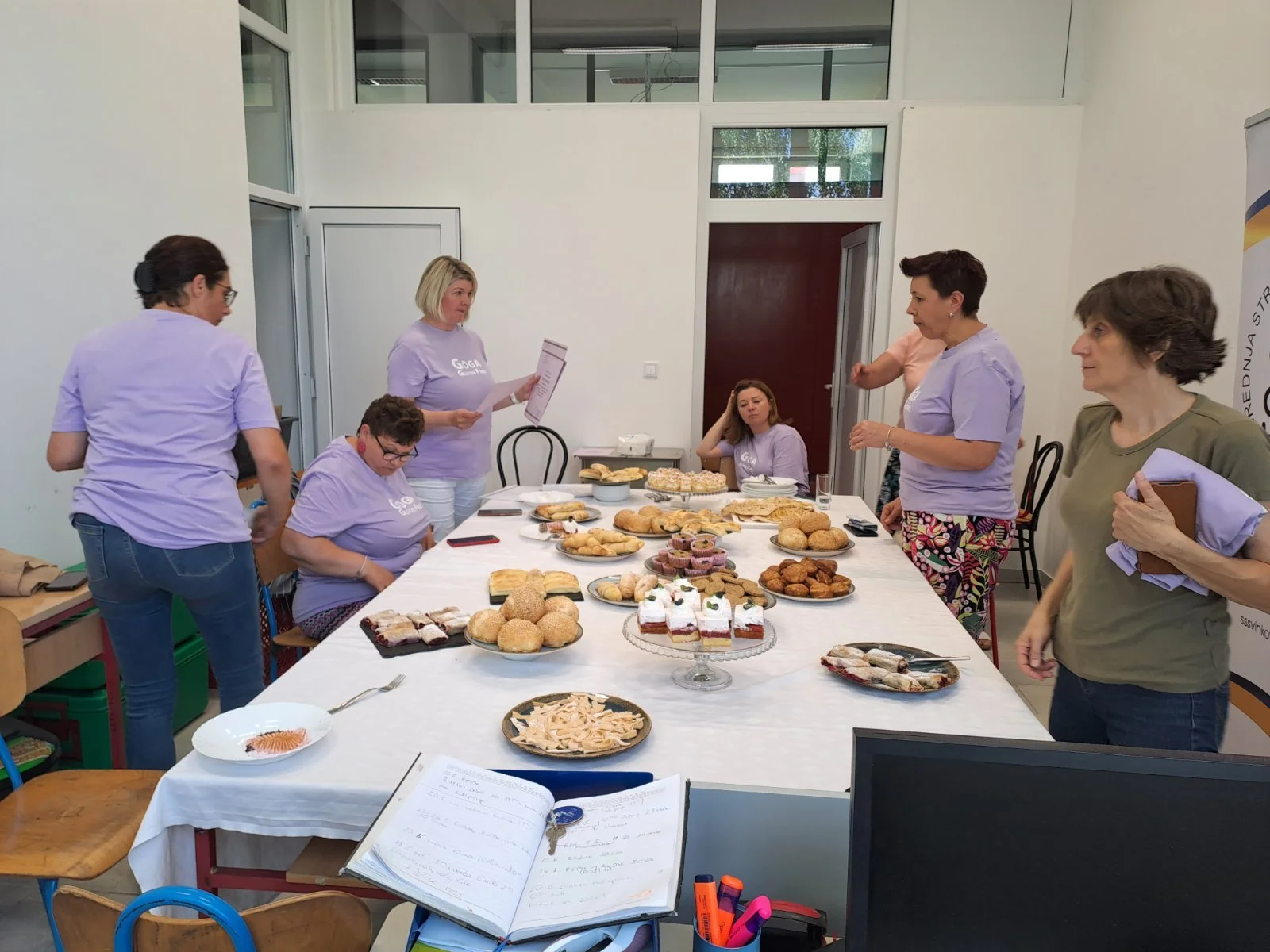 A group of women standing around a table filled with various baked goods, some sitting, in a bright conference room with large windows and white walls.