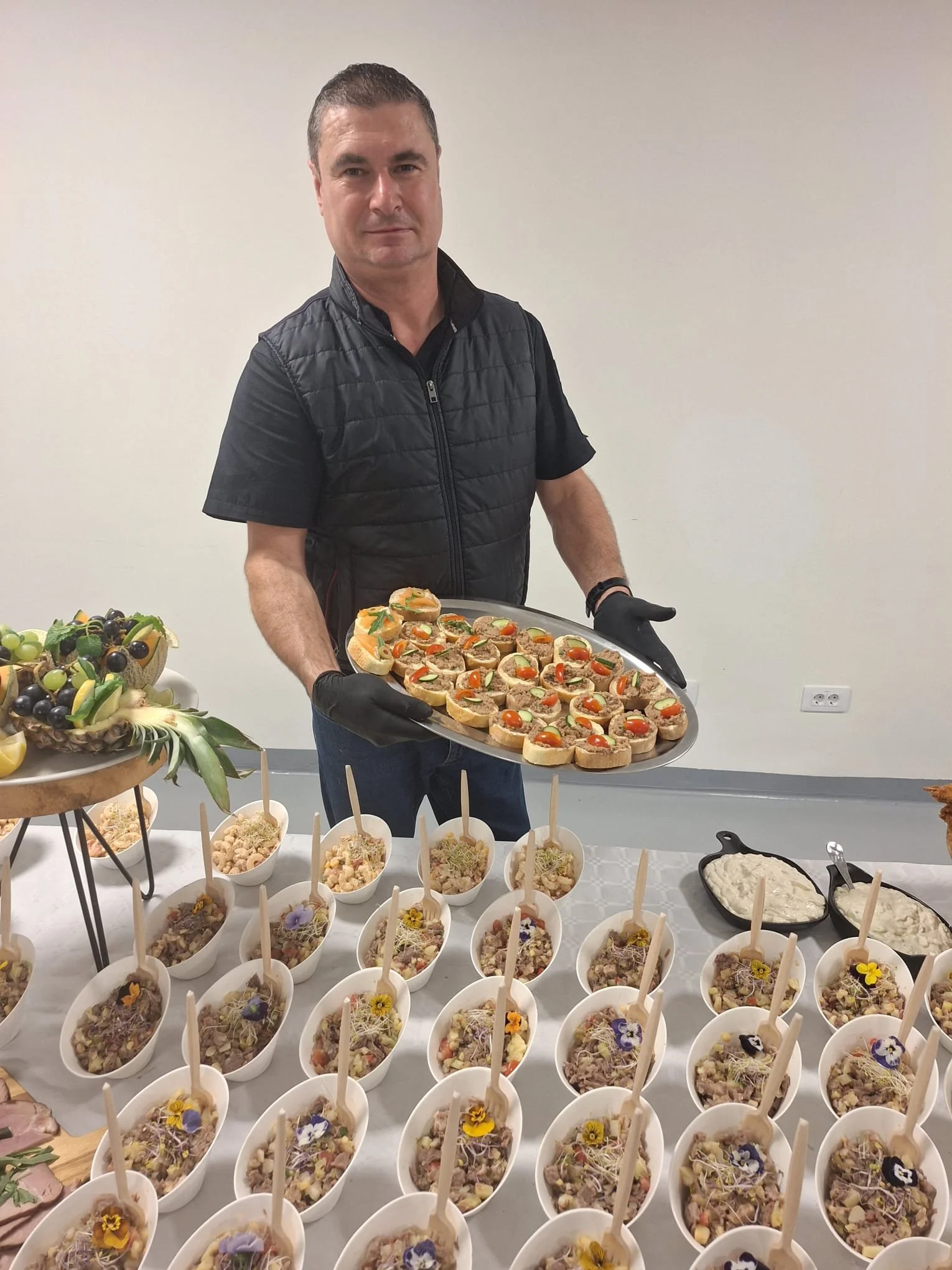 Man in black vest holding a tray of small appetizers at a buffet table with various dishes and fruit in the background.