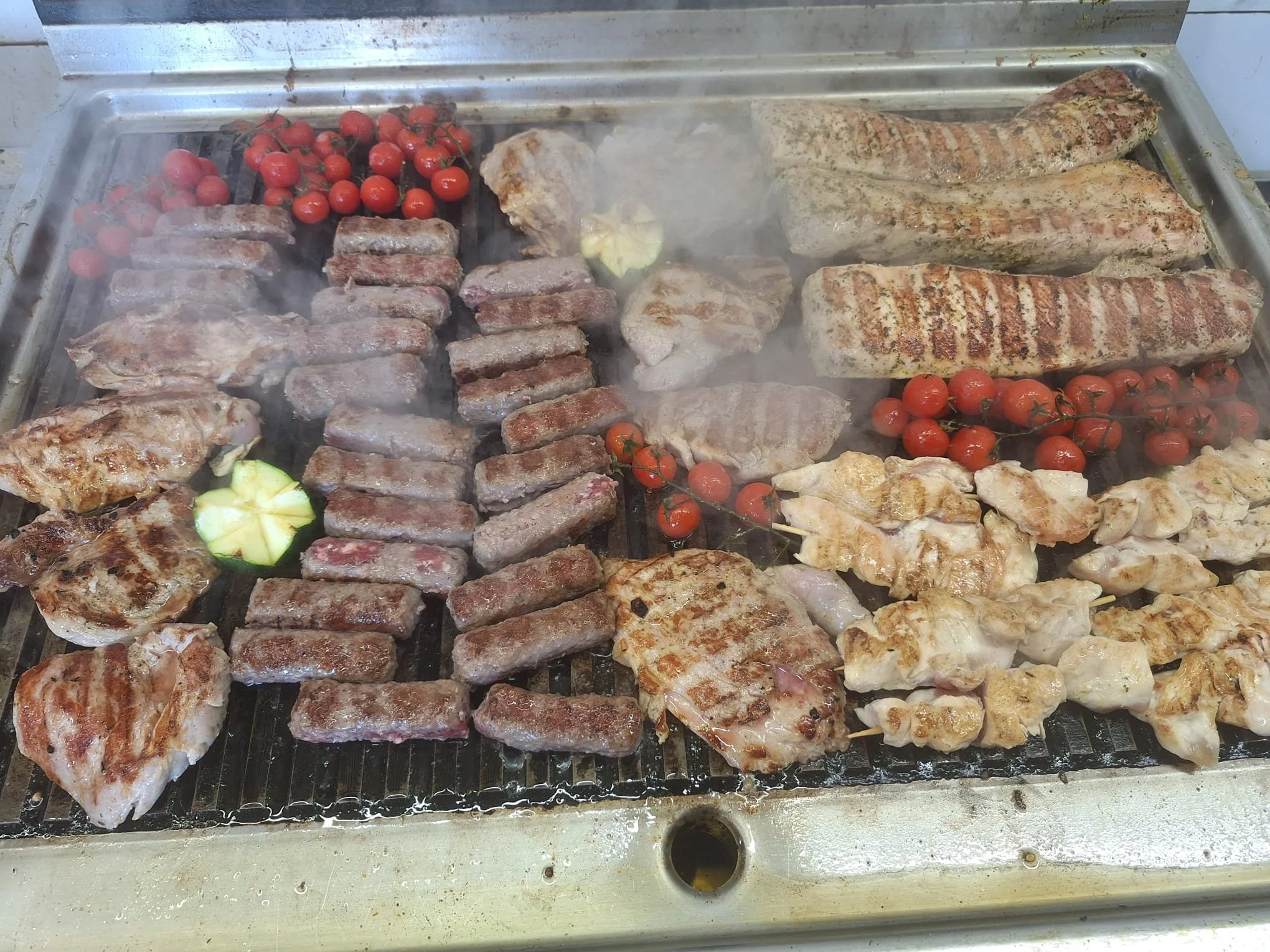 Assorted meats, cherry tomatoes, slices of squash, and skewered chicken grilling on a barbecue grill with smoke.