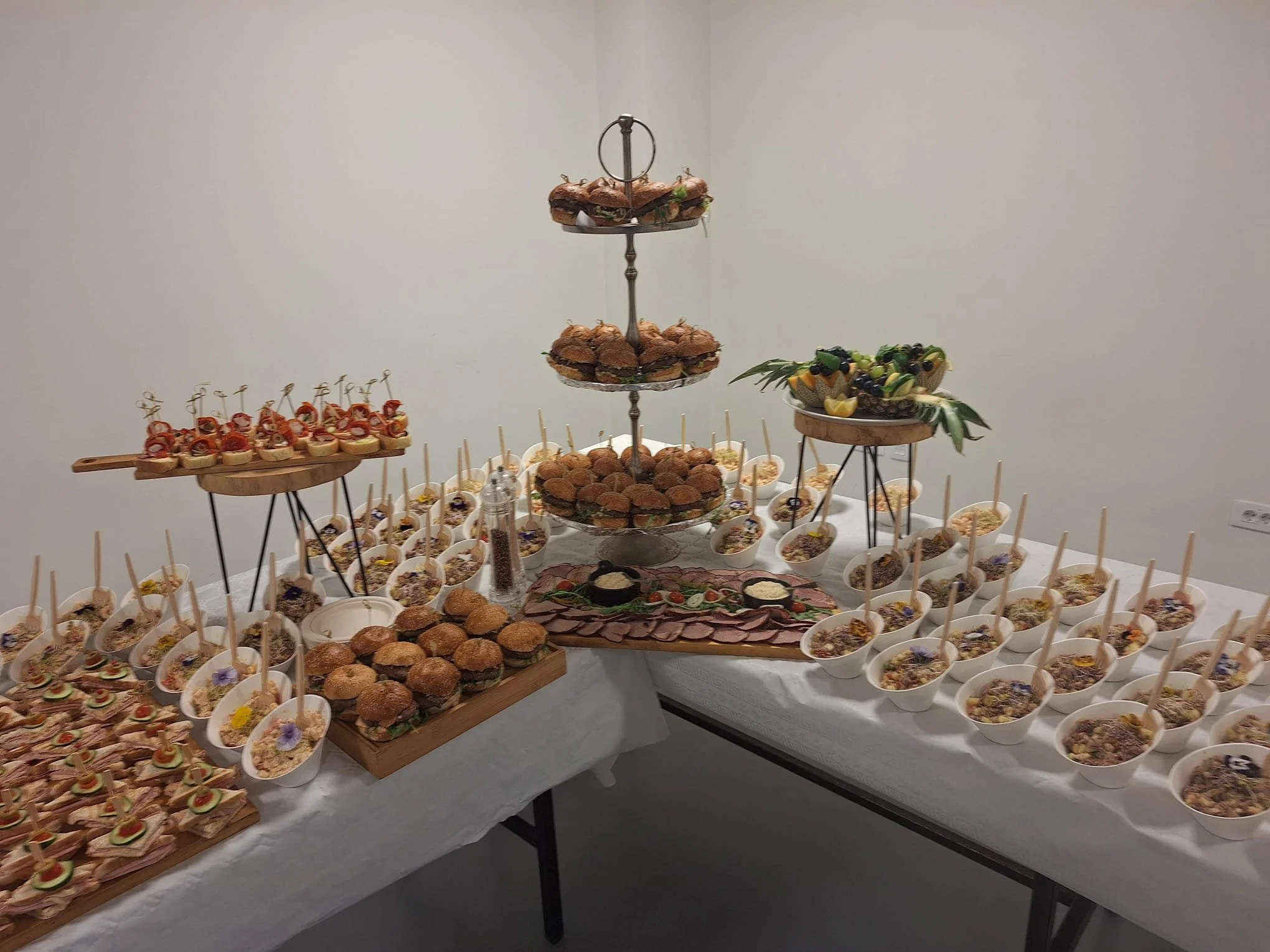 Display of assorted appetizers and finger foods on a buffet table, including mini sandwiches, bowls of salad or dip with spoons, and fruit arrangements.