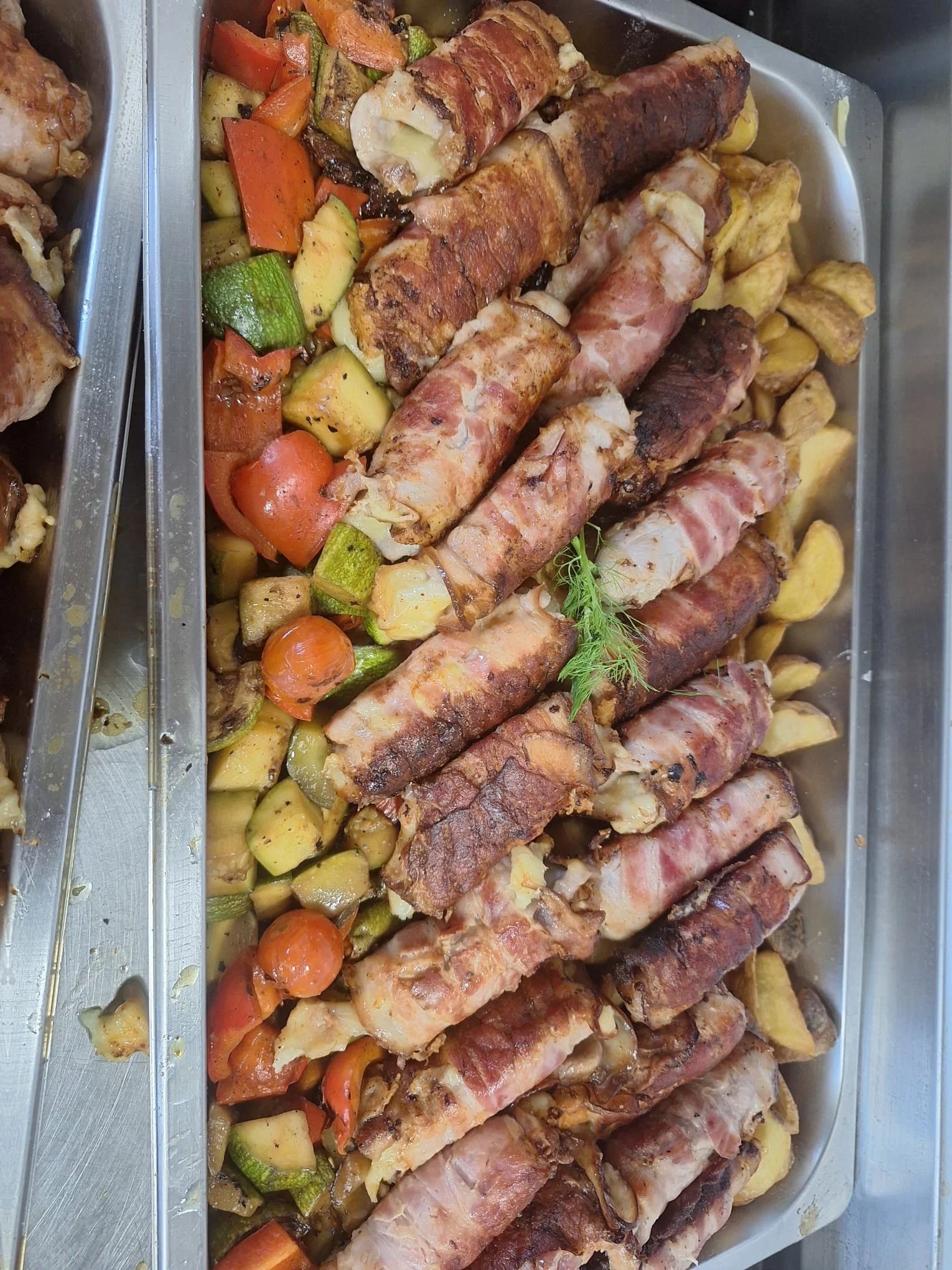 A large tray of cooked bacon-wrapped chicken breasts with a garnish of herbs, accompanied by roasted vegetables including zucchini, tomatoes, carrots, and potatoes.