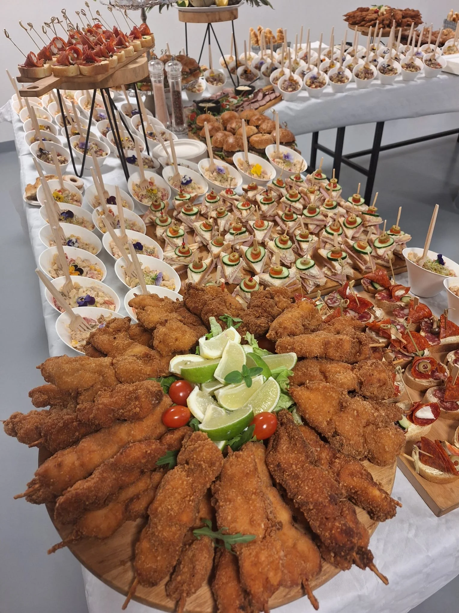A large platter of fried chicken wings garnished with lime wedges, cherry tomatoes, and herbs, with a variety of appetizers and finger foods on the table behind.