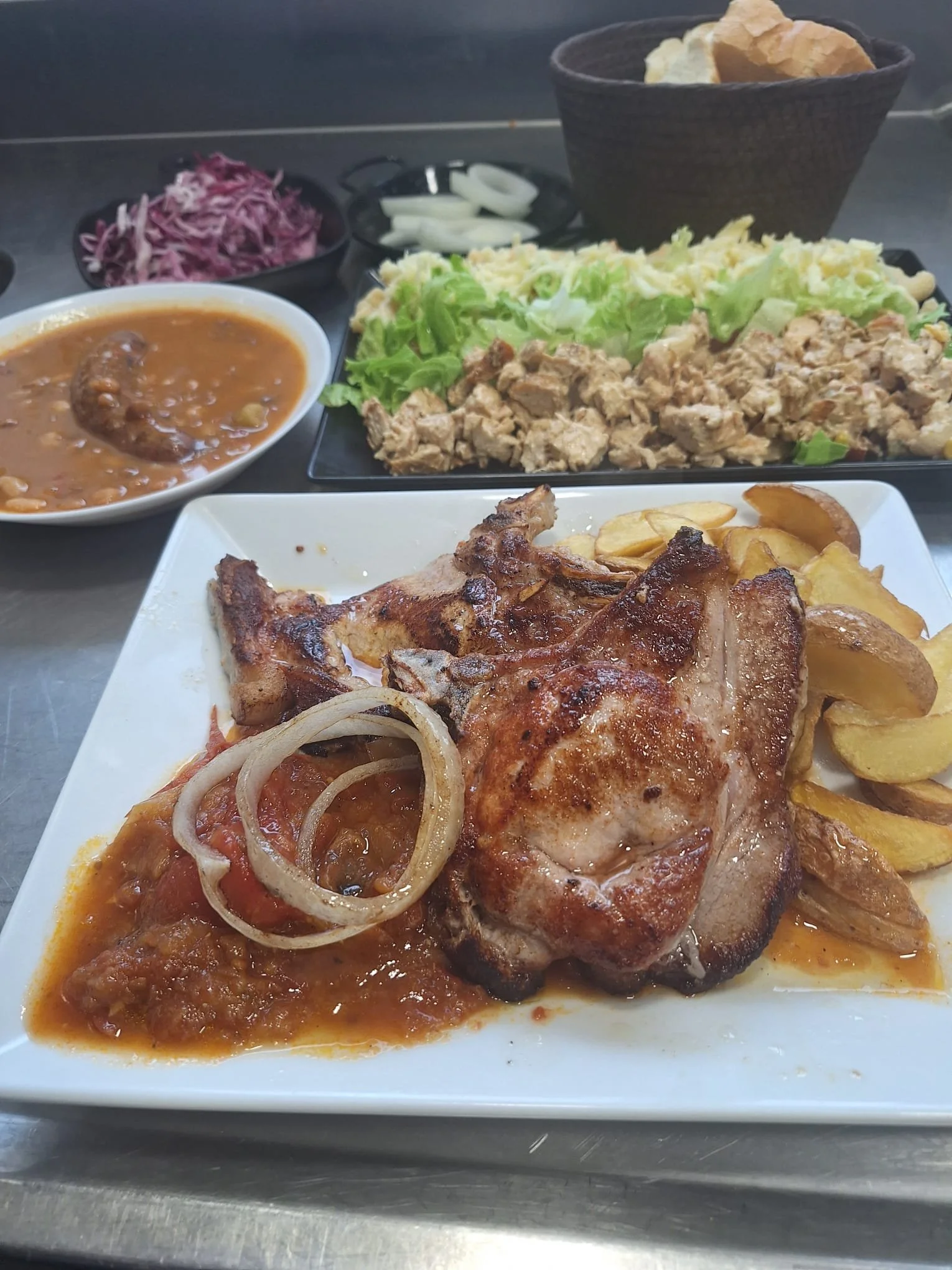 Plate of grilled pork chop with onion rings and potato wedges, side of sauce, salad with lettuce, tomato, and shredded cheese, rice, black beans, shredded red cabbage, sliced onions, and a bowl of potato chips.