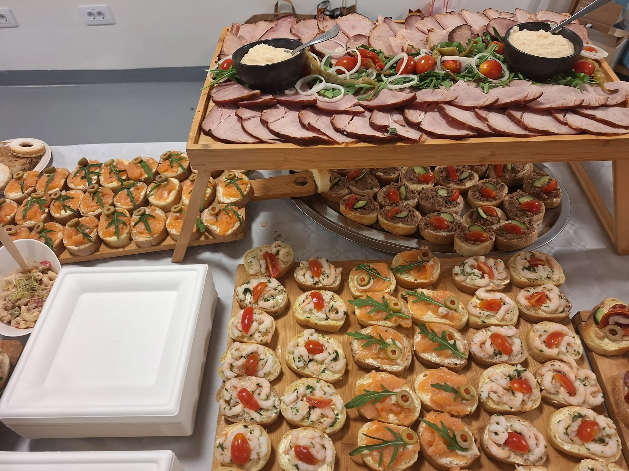 Assorted appetizers including sliced meats, small canapés with seafood, vegetables, and cheese, arranged on wooden and metal trays, with some garnished with cherry tomatoes and herbs, on a white tablecloth.