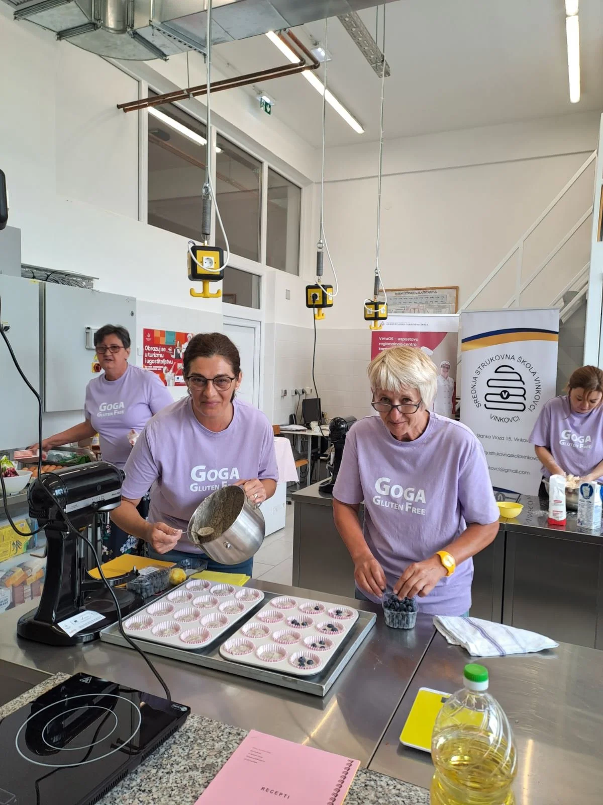 Four women in lavender t-shirts labeled 'GOGA Gluten Free' preparing blueberry muffins in a commercial kitchen.