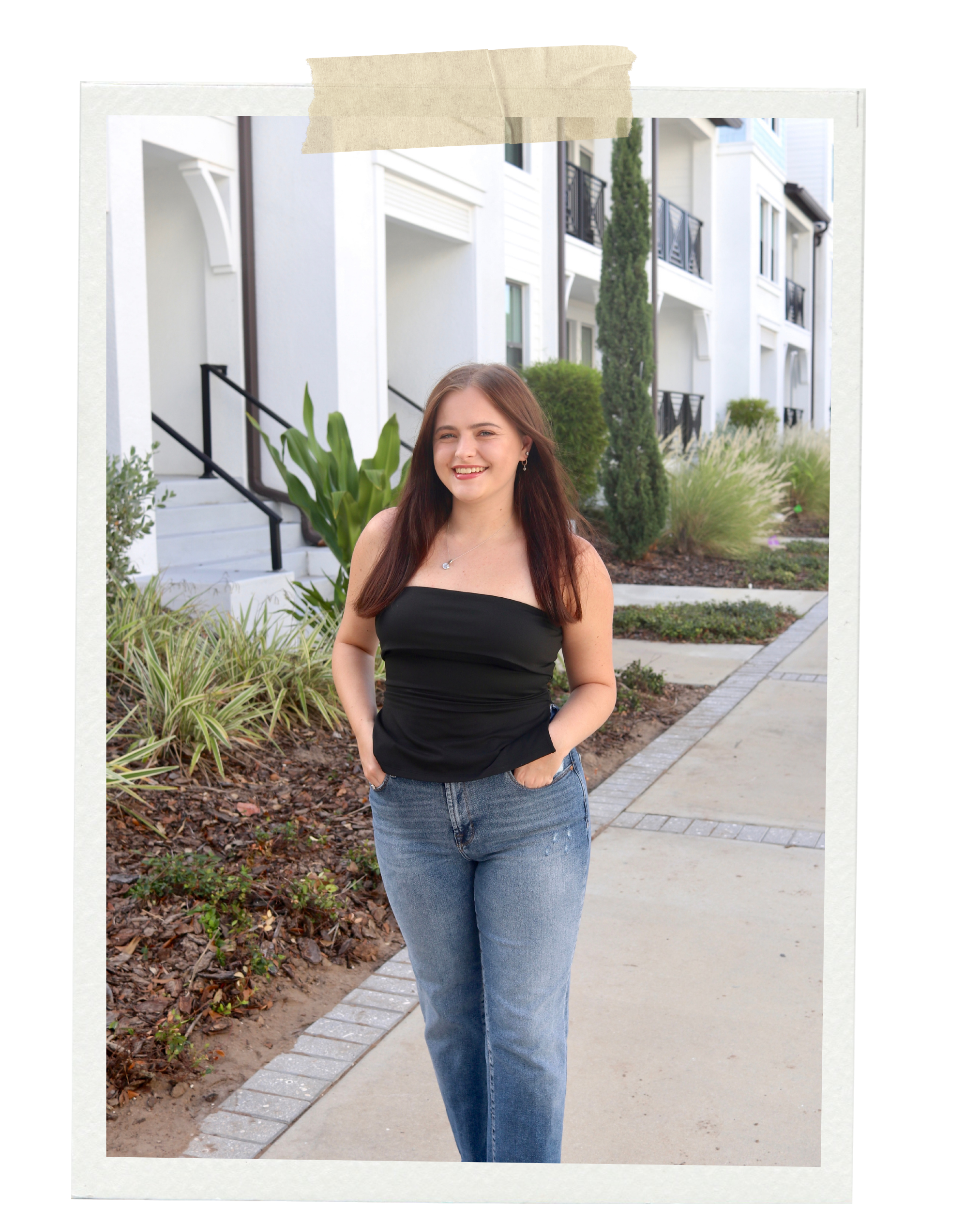 Young woman with long brown hair smiling, standing outdoors on a sidewalk in front of green plants and white apartment buildings.
