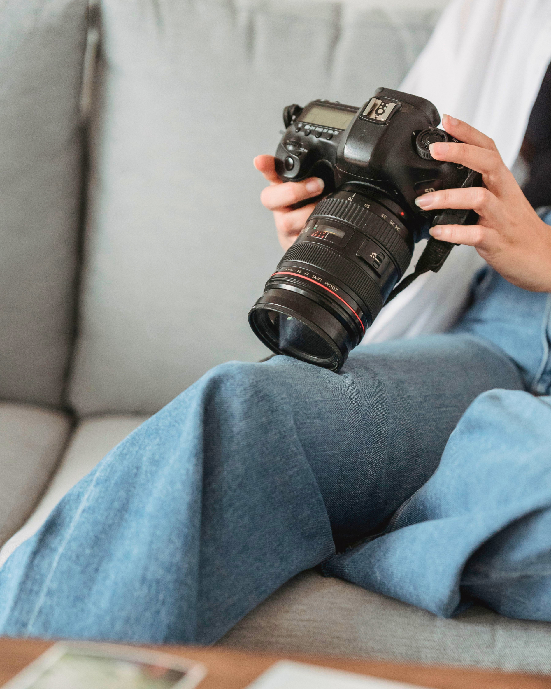 A person holding a black professional camera with a large zoom lens, seated on a beige sofa with grey pants visible.