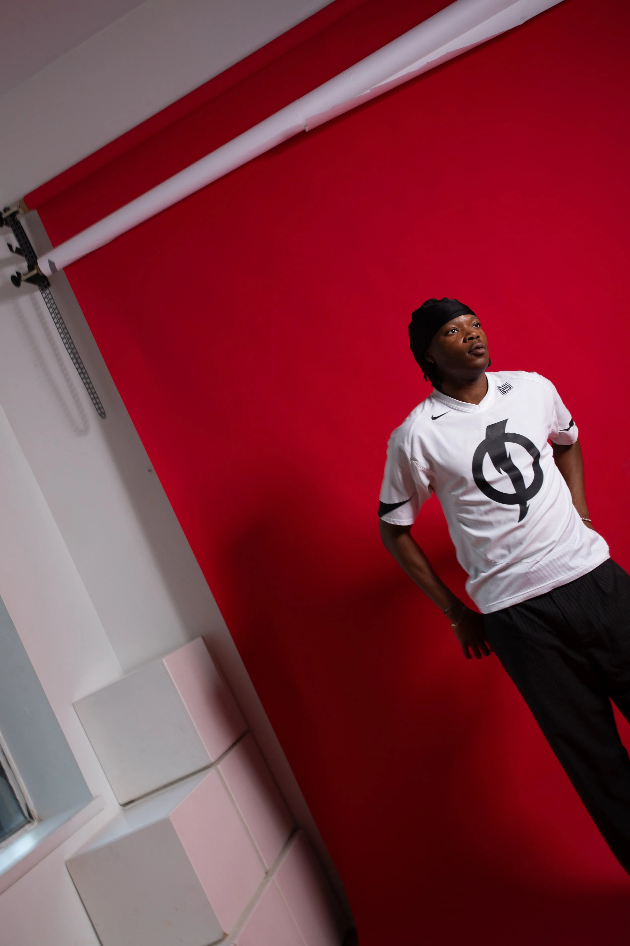 Person posing in front of a red backdrop, wearing a white sports jersey with a black logo and black pants.