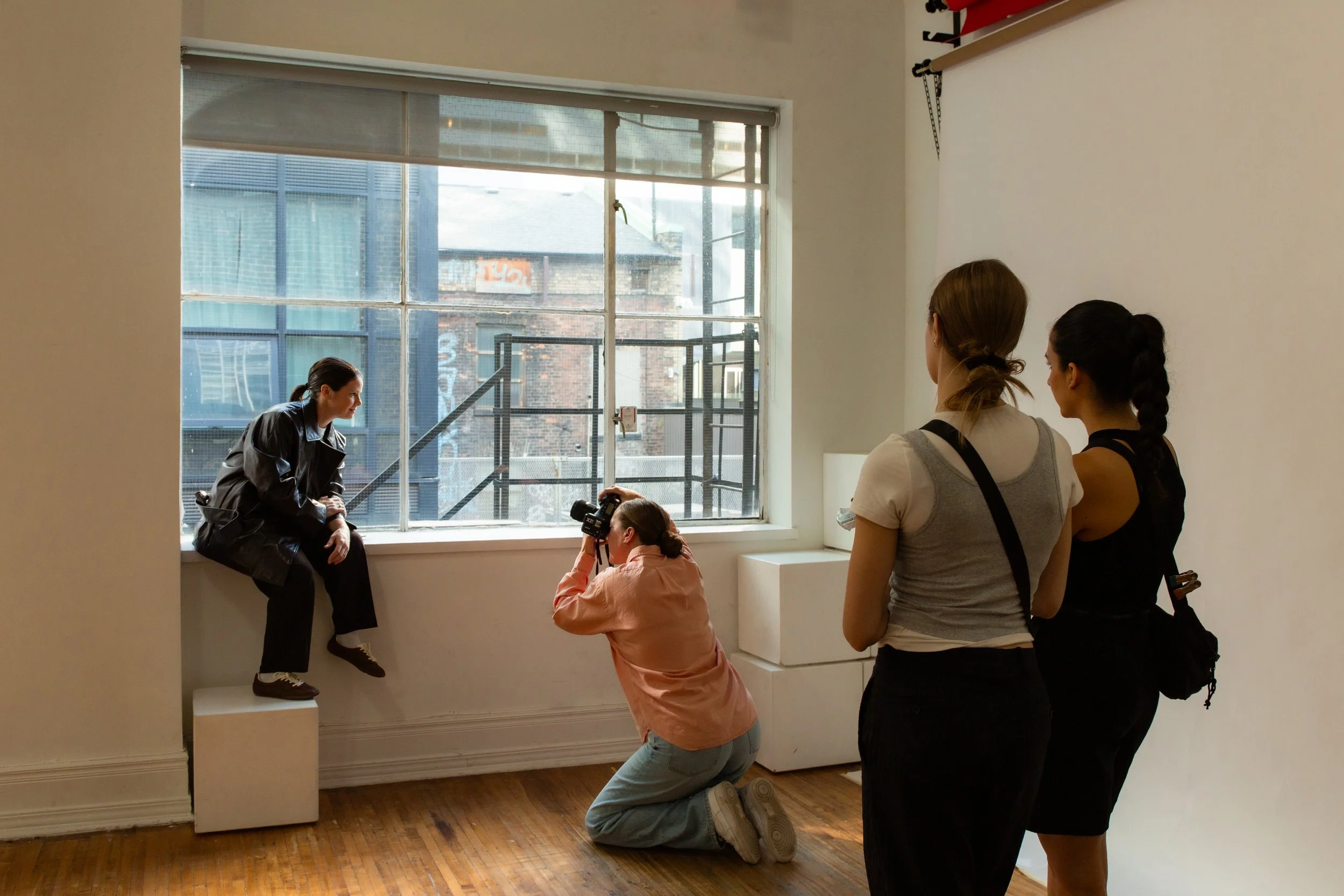 A photoshoot in a studio features a woman sitting on a ledge near a large window, with a photographer kneeling taking her picture. Two women stand observing, one with a gray shirt and another with a black top. The room has wooden floors and white walls.