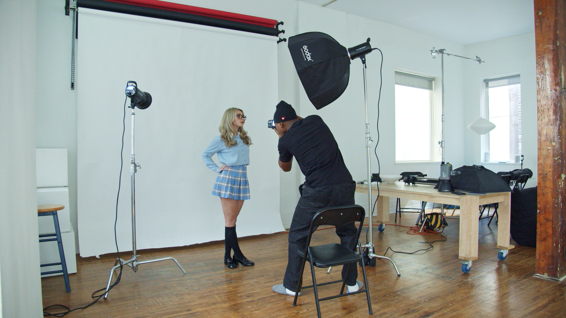 A woman wearing glasses and a blue plaid skirt poses for a photo in a photography studio with a white backdrop, while a photographer takes her picture using professional lighting equipment.