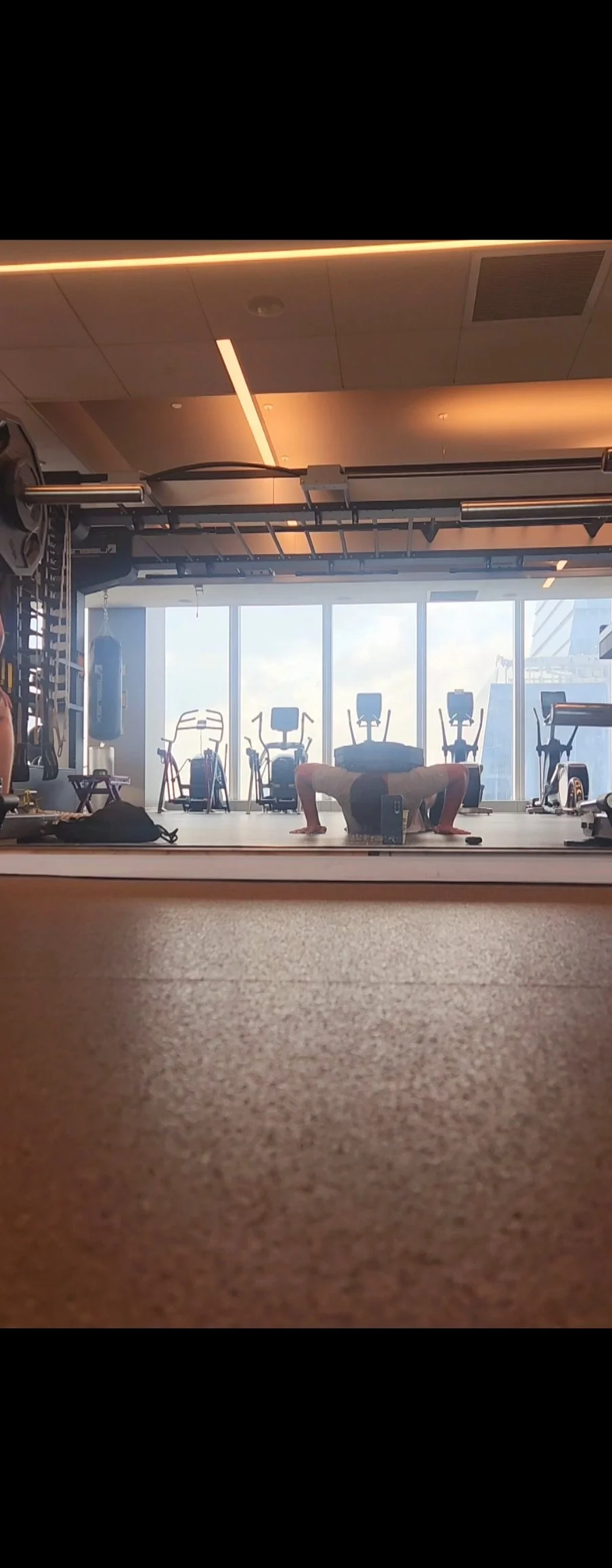 A person doing a push-up in a gym with workout equipment and large windows in the background.