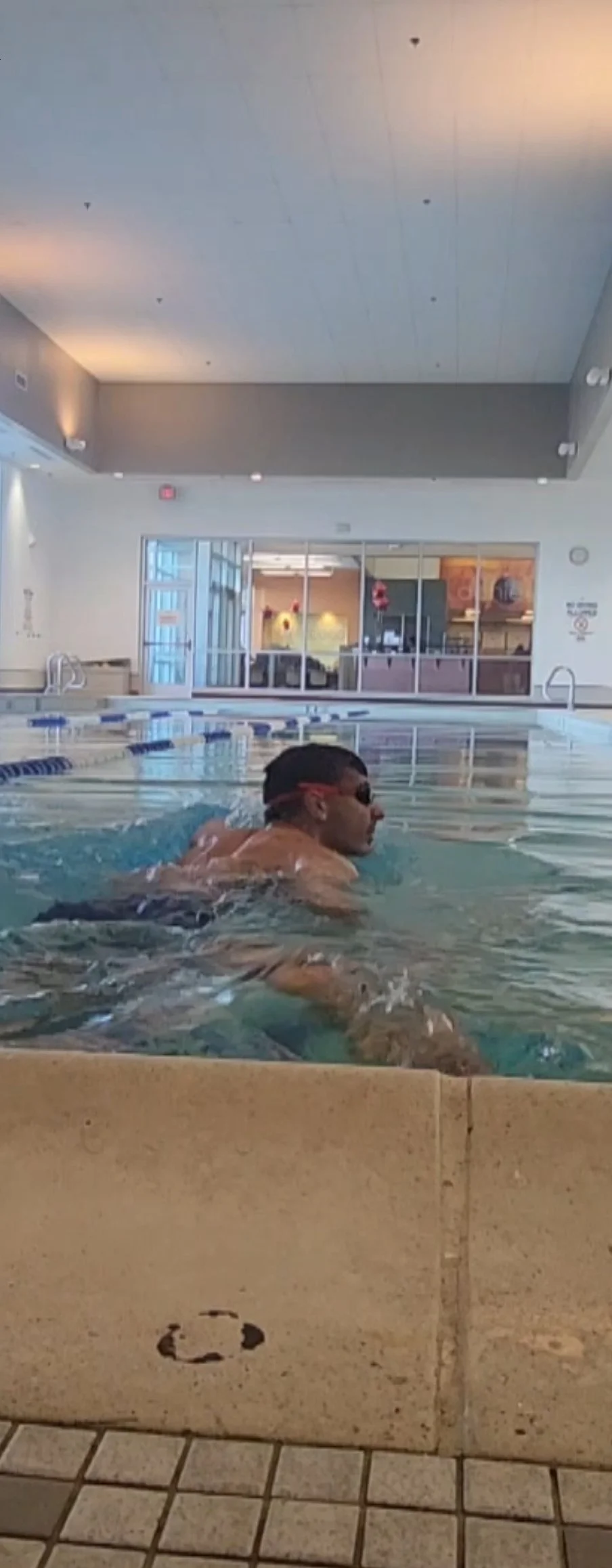 Man wearing goggles swimming in an indoor pool with a lounging area and glass window in the background.