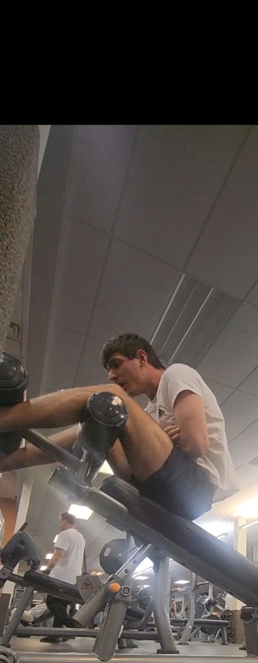 A young man in a white T-shirt is sitting on an inclined back extension bench at a gym, leaning forward and engaging in a workout. There is gym equipment visible around him, and another person is working out in the background.