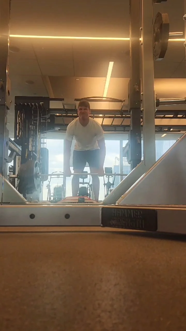 A man lifting a barbell in a gym, seen through a mirror reflection, with gym equipment and large windows in the background.