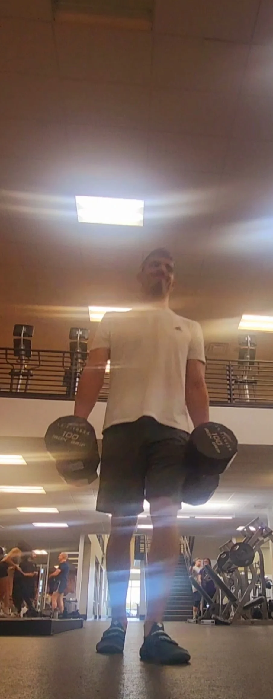 A man in a white t-shirt and black shorts holding dumbbells in a gym. The photo is taken from a low angle showing the ceiling and gym equipment in the background.