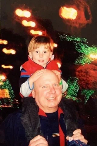 A young boy sitting on an adult man's shoulders at night, with holiday lights in the background, smiling at the camera.