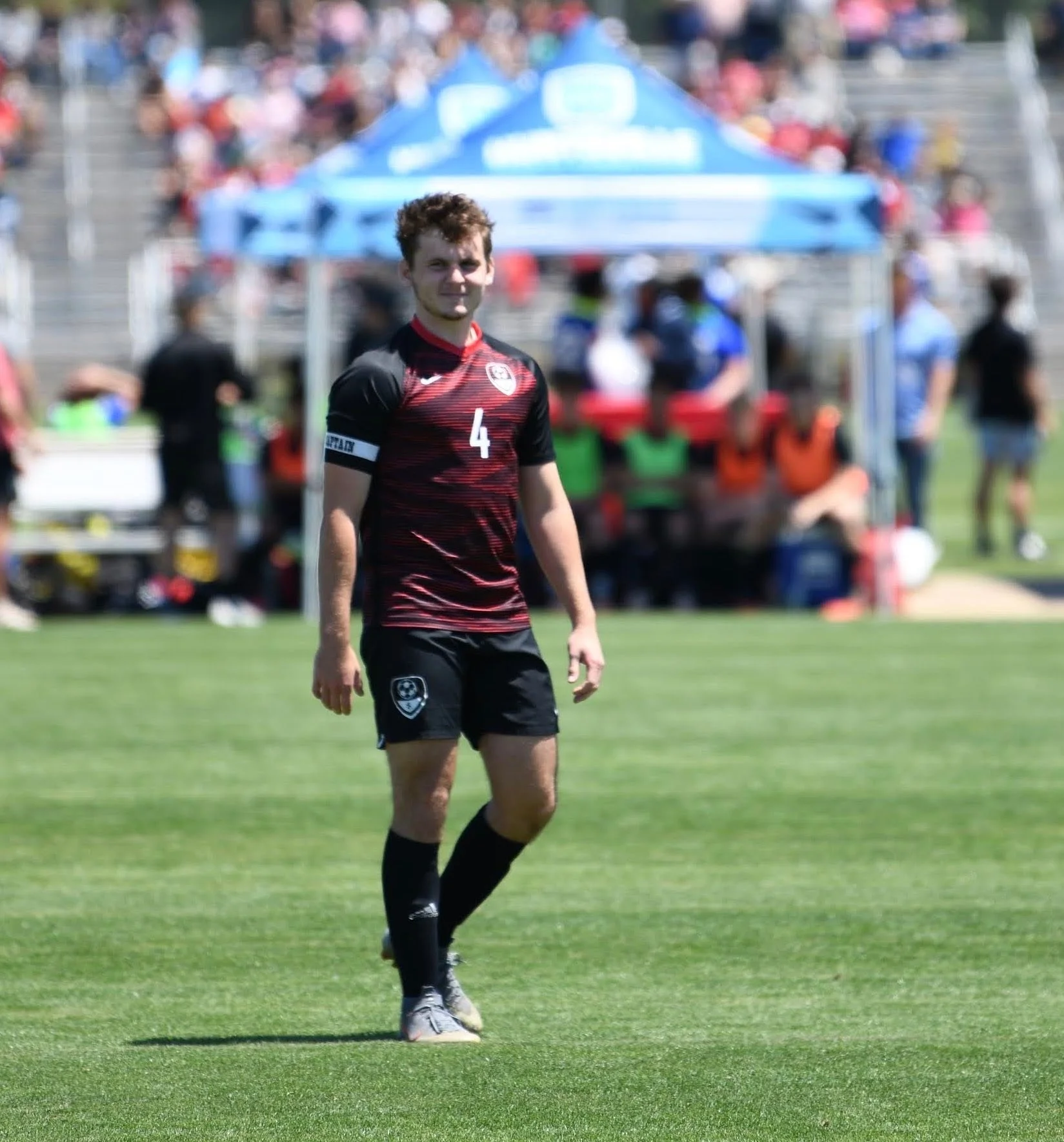 A male soccer player on the field wearing a black and red jersey with the number 4, black shorts, and black socks, standing on green grass during a game or practice, with spectators and people sitting under a blue tent in the background.