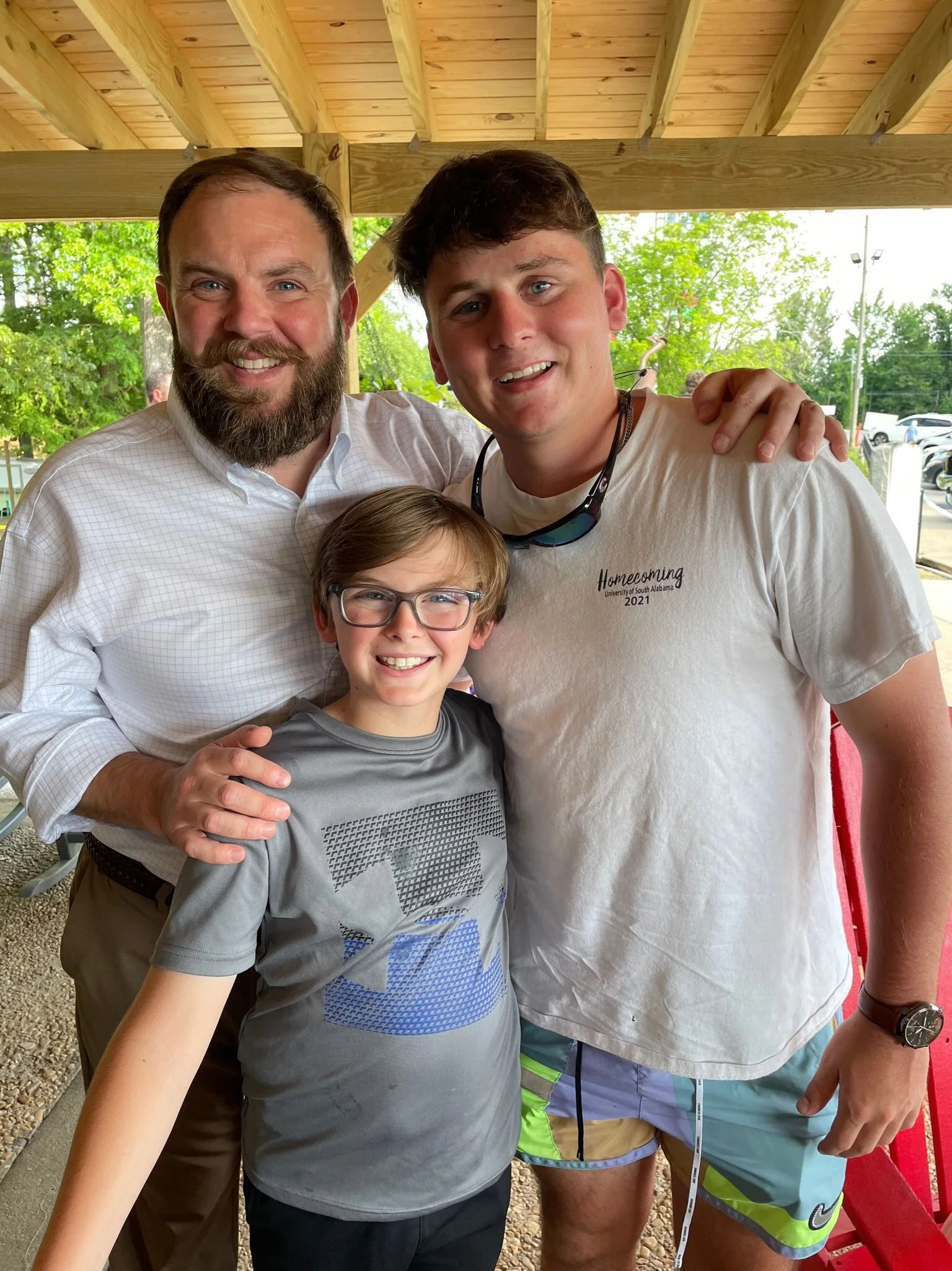 Three people smiling, a man with a beard on the left, a teenage boy on the right, and a young boy with glasses in the middle, standing under a wooden shelter with green trees and parking lot in the background.