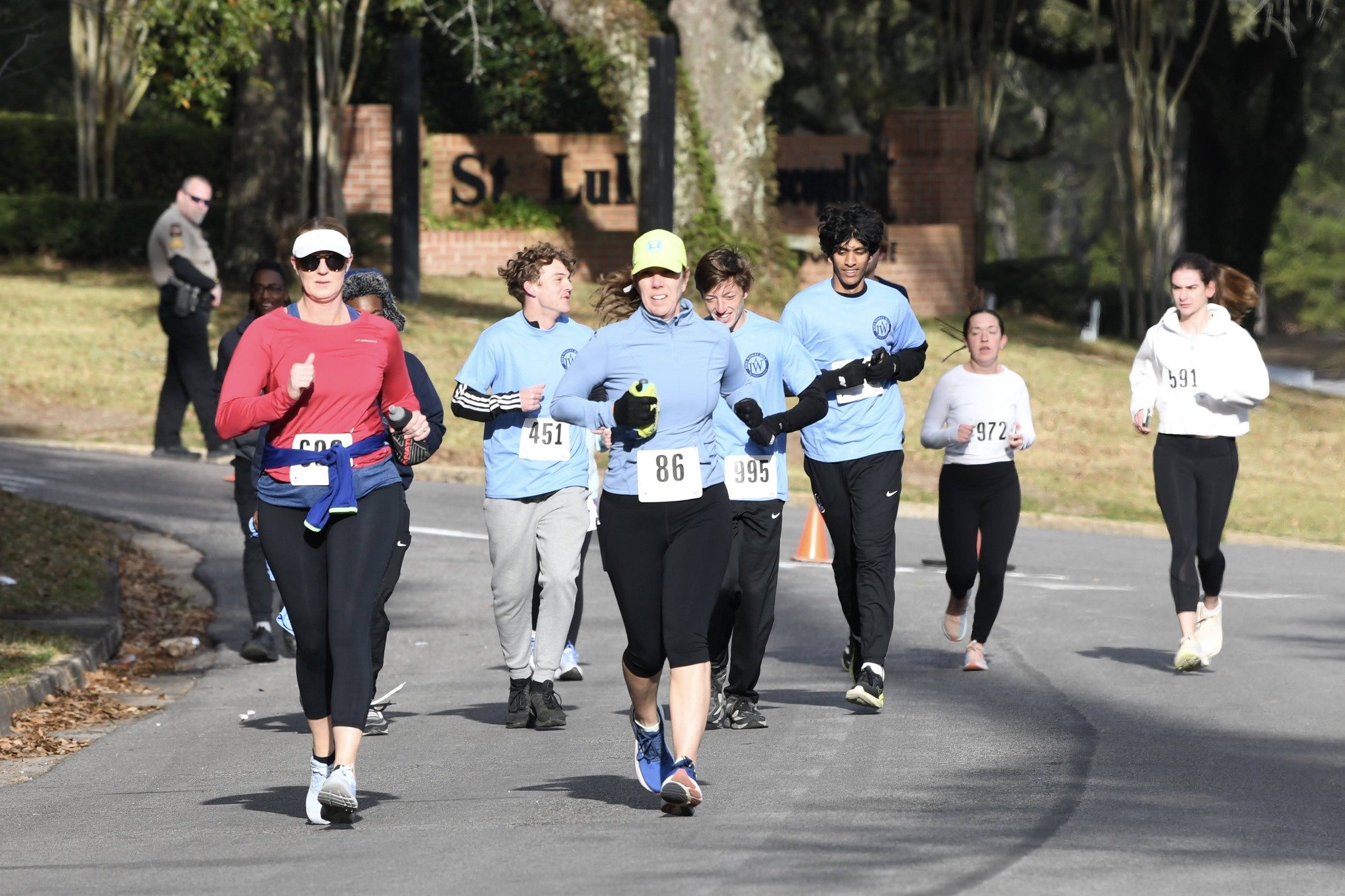 Group of runners participating in a race, running on a street. They wear race bibs, athletic clothing, and are outdoors with trees and a brick sign in the background.