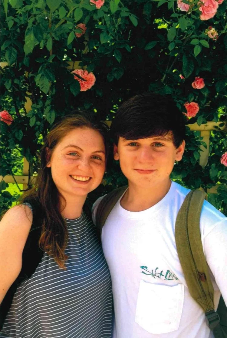 A young woman and young man taking a selfie outside in front of a bush with pink flowers, smiling and wearing casual clothes and backpacks.