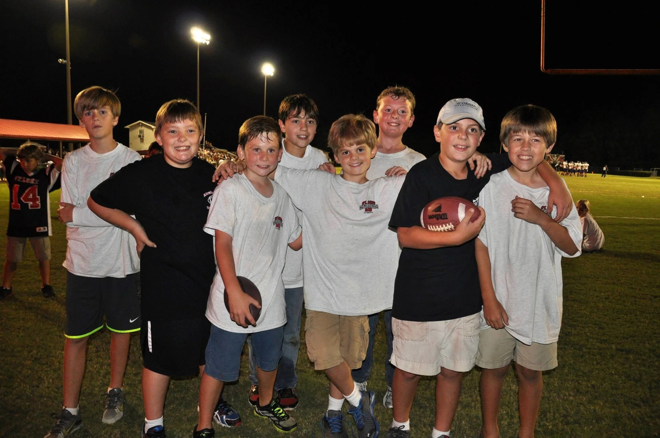 Group of young boys at night on a football field, some holding footballs, posing for a photo, stadium lights overhead, football game in the background.