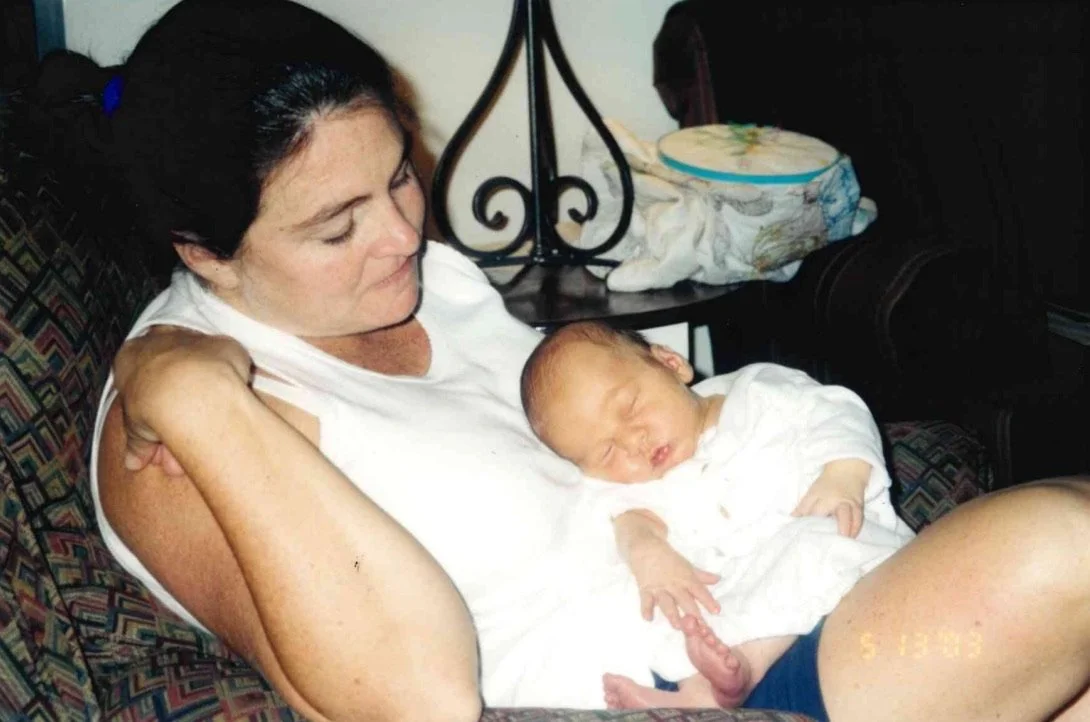 A woman with dark hair holding a sleeping newborn baby on her lap, sitting on a patterned couch in a cozy room.