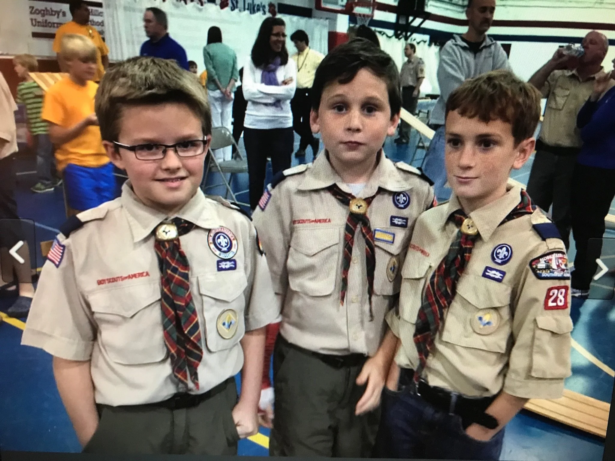 Three young boys dressed in Cub Scout uniforms standing close together at a gathering in a gymnasium. They are surrounded by other people, some standing and some walking, with tables and chairs visible in the background.