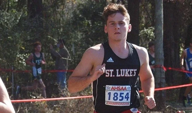 Young male runner wearing a black St. Luke's uniform and bib number 1854 competing in a cross-country race outdoors with a wooded background.