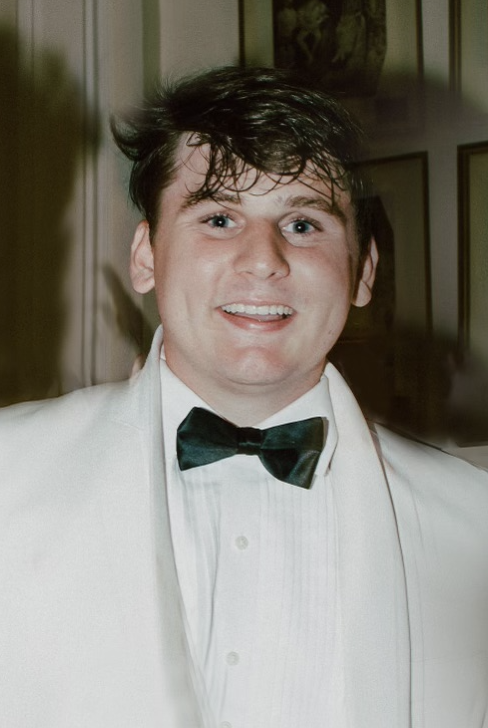 Young man wearing a white tuxedo with a black bow tie, smiling, with slightly messy dark hair, indoors with a painting or photograph in the background.