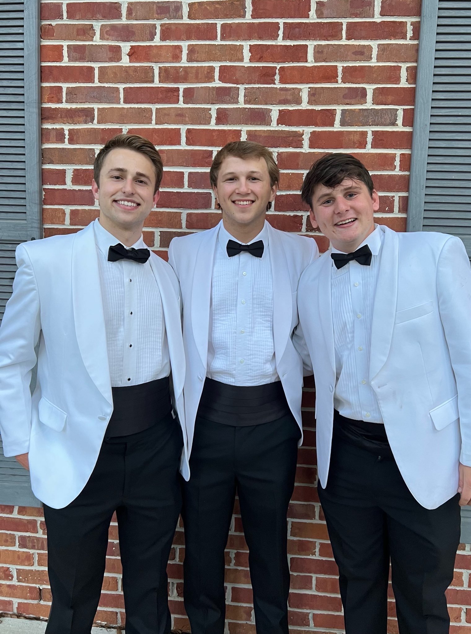 Three young men wearing white tuxedos with black bow ties and black pants, standing in front of a brick wall with gray window shutters, smiling at the camera.