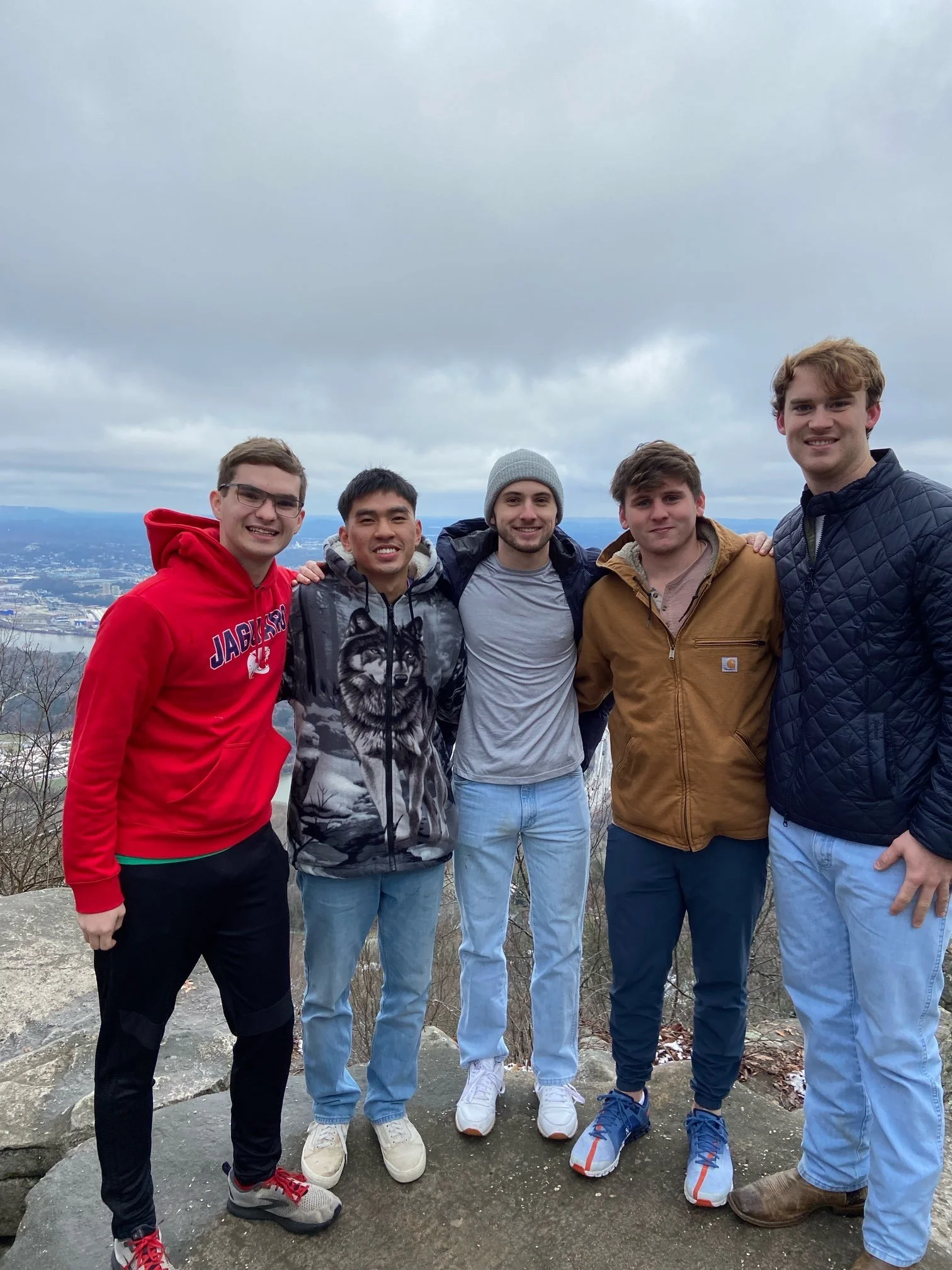 Five young men standing together outdoors on a cloudy day, smiling, with a view of a city and mountains in the background.