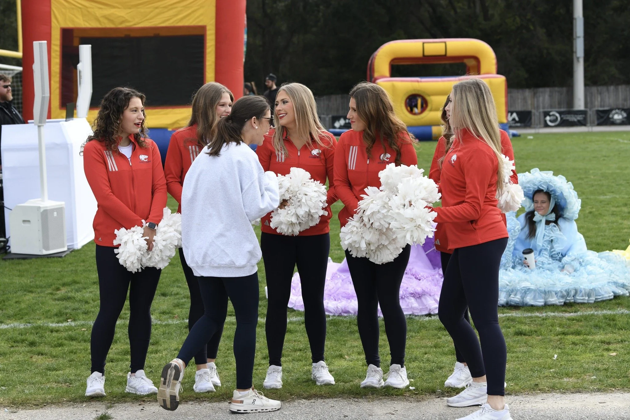 Group of young women in matching red jackets and black leggings holding white pom-poms, standing on a grassy field during an outdoor event, with a girl in a blue costume sitting on the ground in the background.